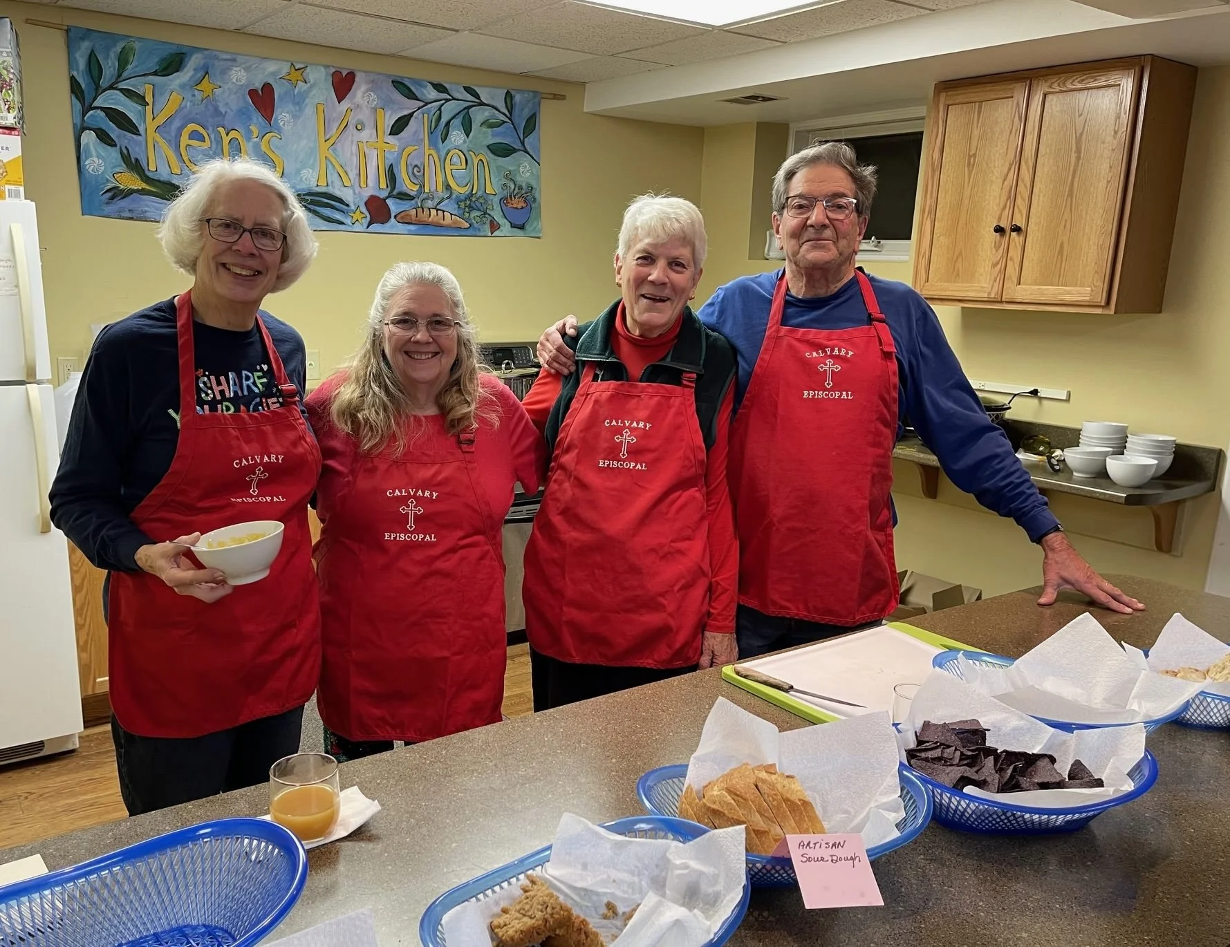 Three woman and one man wearing red aprons standing behind the kitchen counter with baskets of bread. A sign reading “Ken’s Kitchen” is behind them.