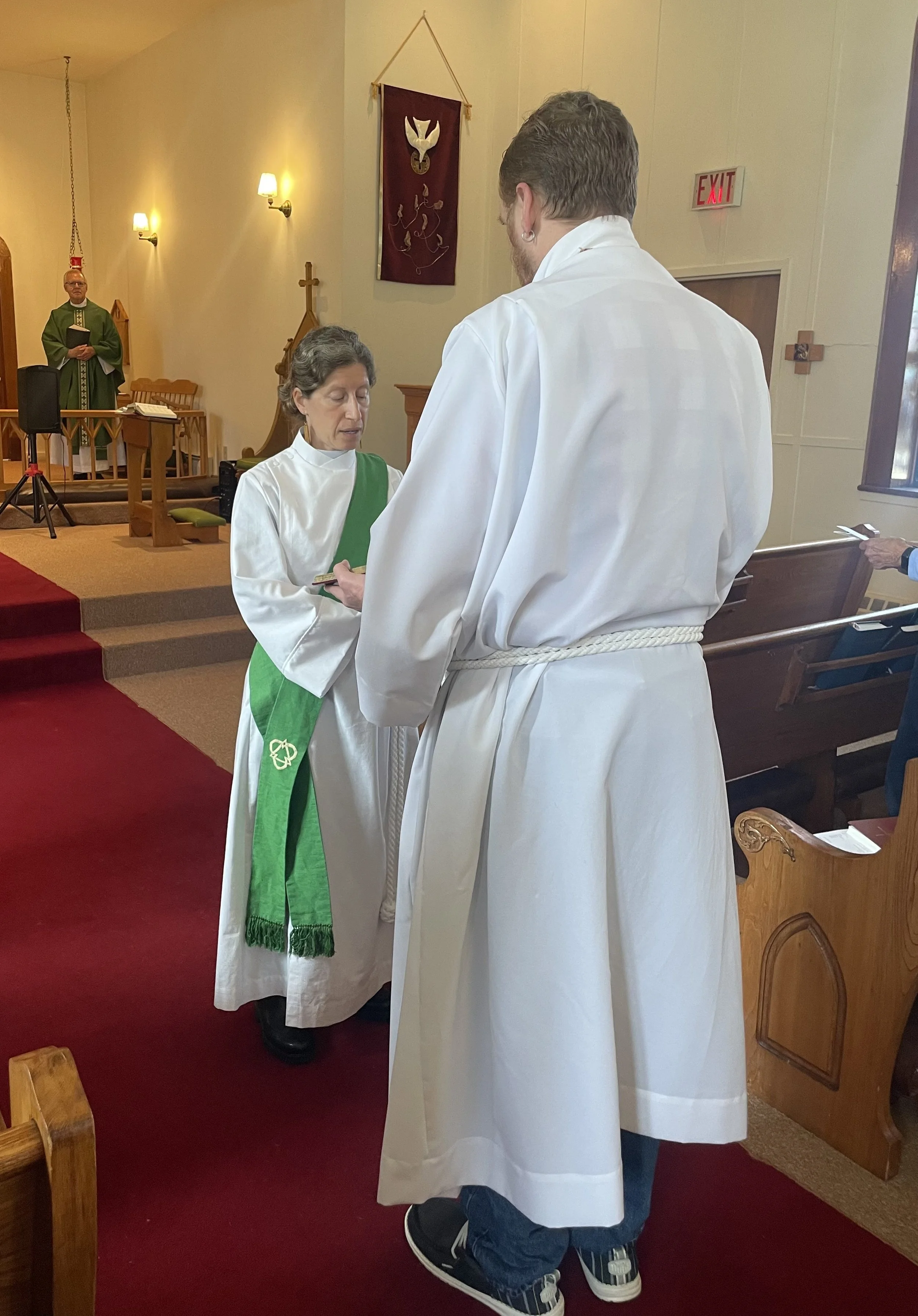 A church ceremony with a woman priest and a man standing in front of the altar. The priest wears a white robe with a green stole and is reading from the Bible and the man wears a white robe and is holding the Bible. Another priest stands behind them.