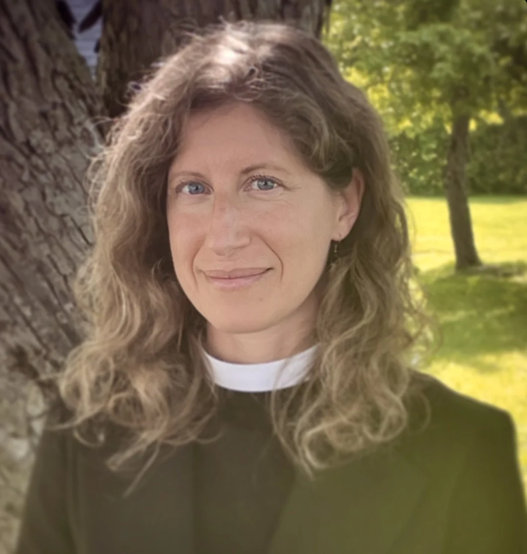 A woman with wavy, shoulder-length hair, a gentle smile, and a clerical collar standing outdoors beside a tree with green leaves in the background.