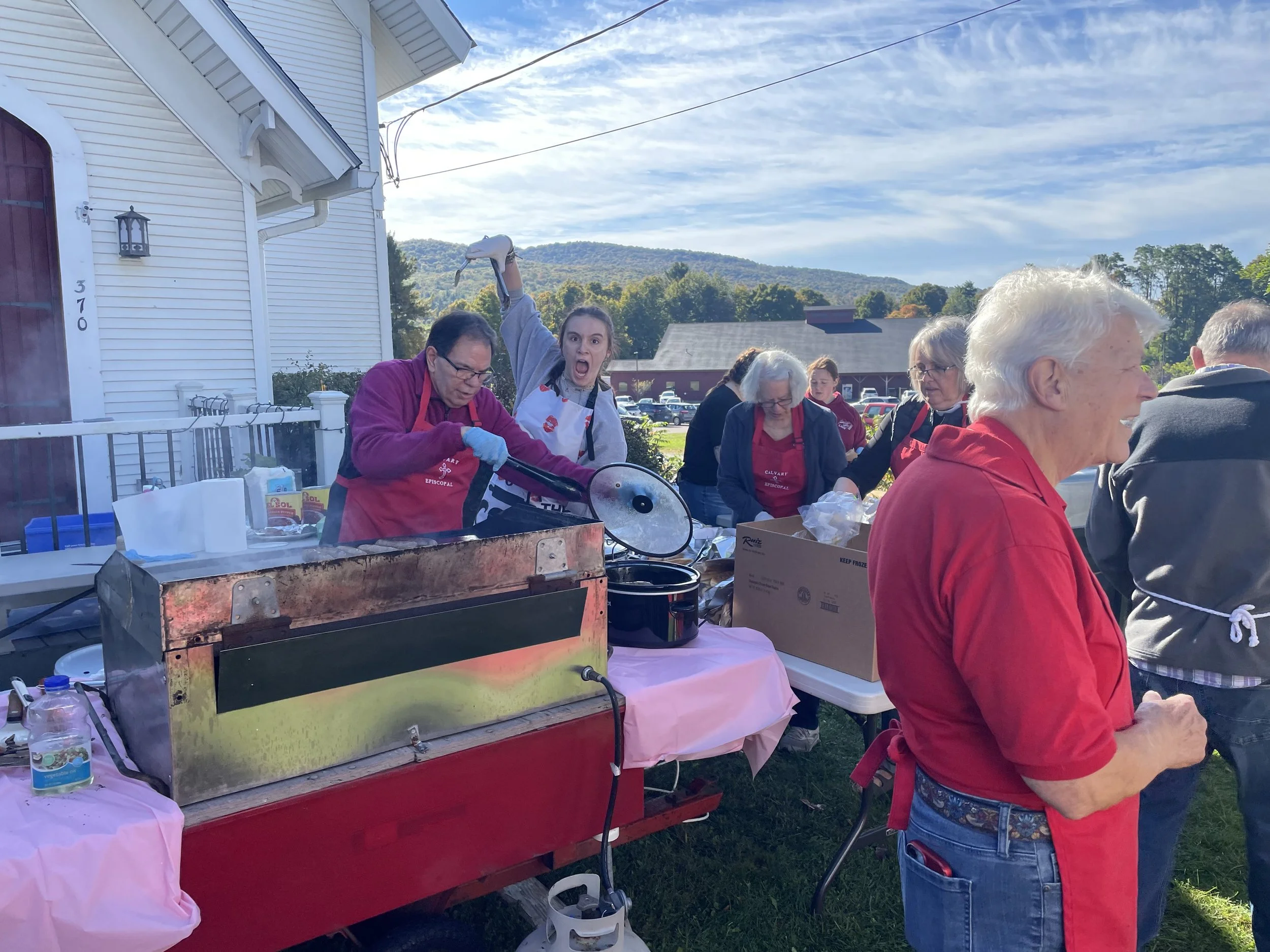Man and young woman behind grill at the Calvary sausage and peppers stand. The woman is making a funny face holding grilling tongs over the man’s head.