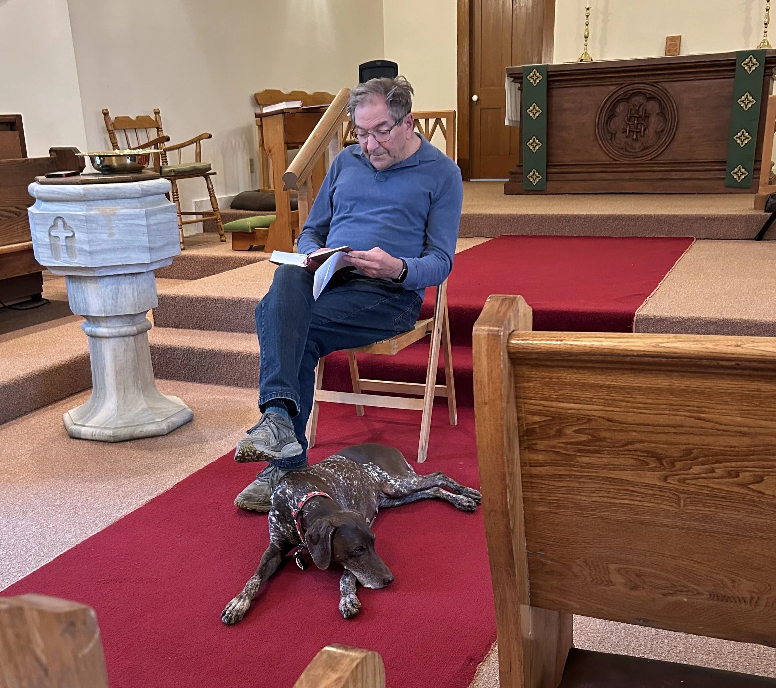 Man in blue shirt and blue jeans reading a prayer book with brown dog resting at his feet.