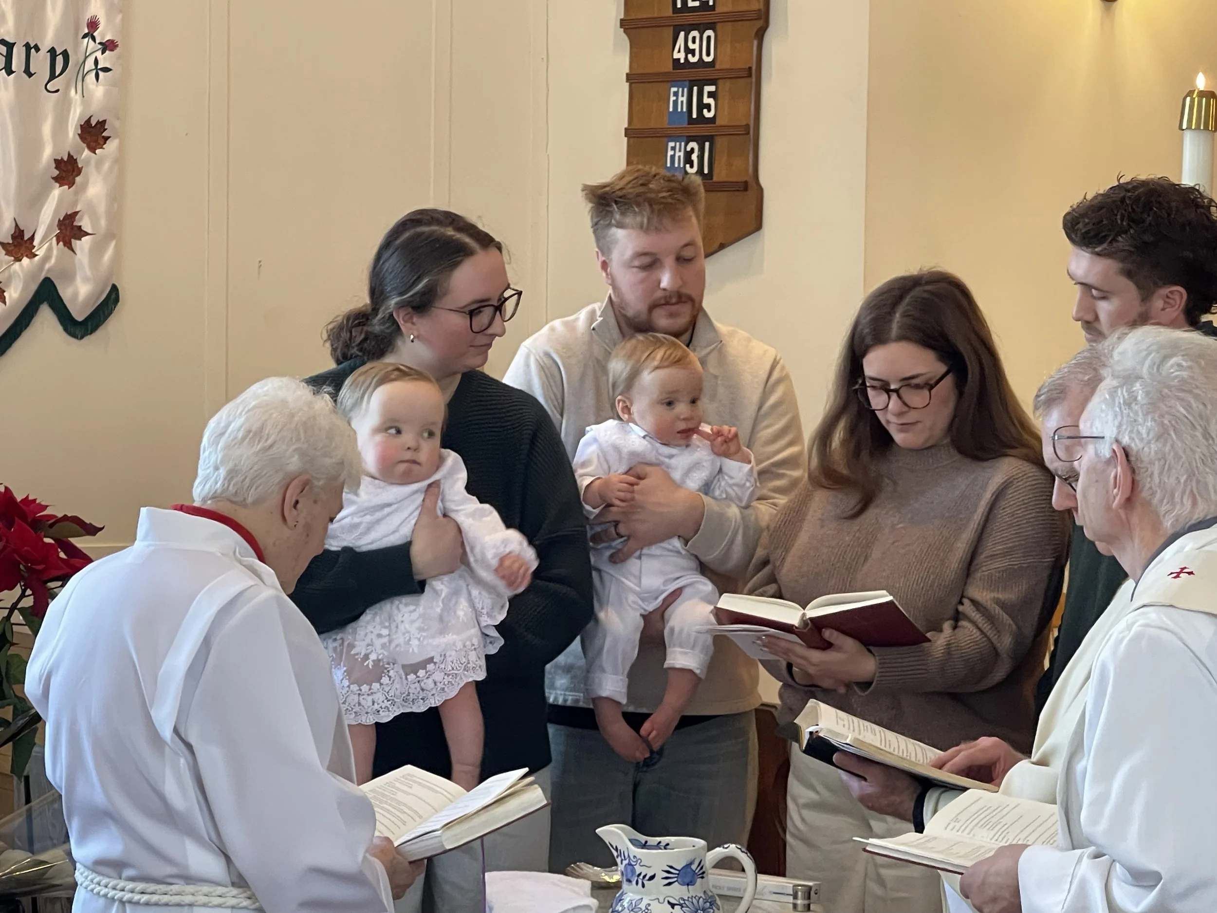 A baptism ceremony with twin babies, Mom, dad, godparents, and three clergy members surrounding the baptismal font.