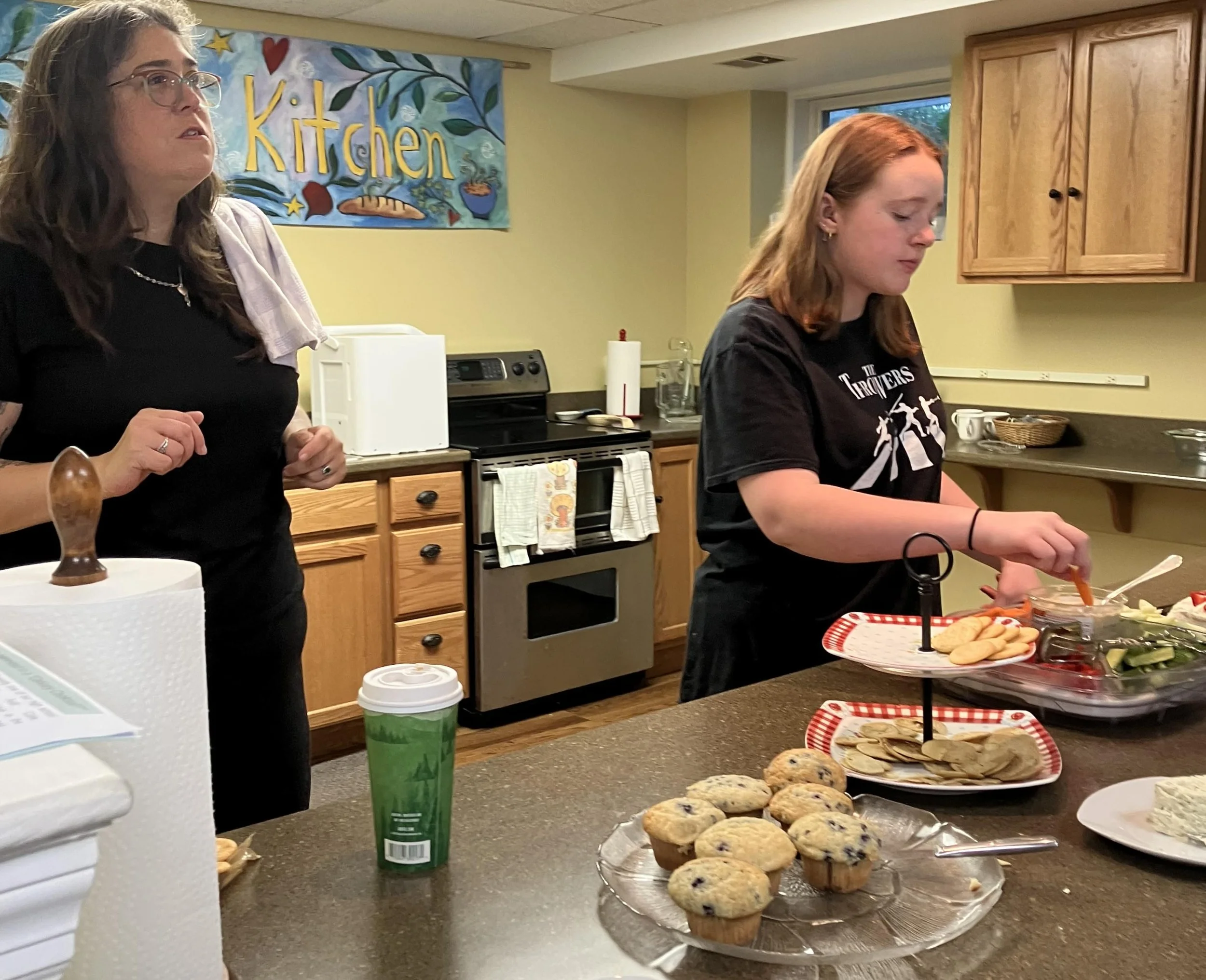 Mother and daughter serving food at kitchen counter during coffee hour with muffins, cheese and crackers.