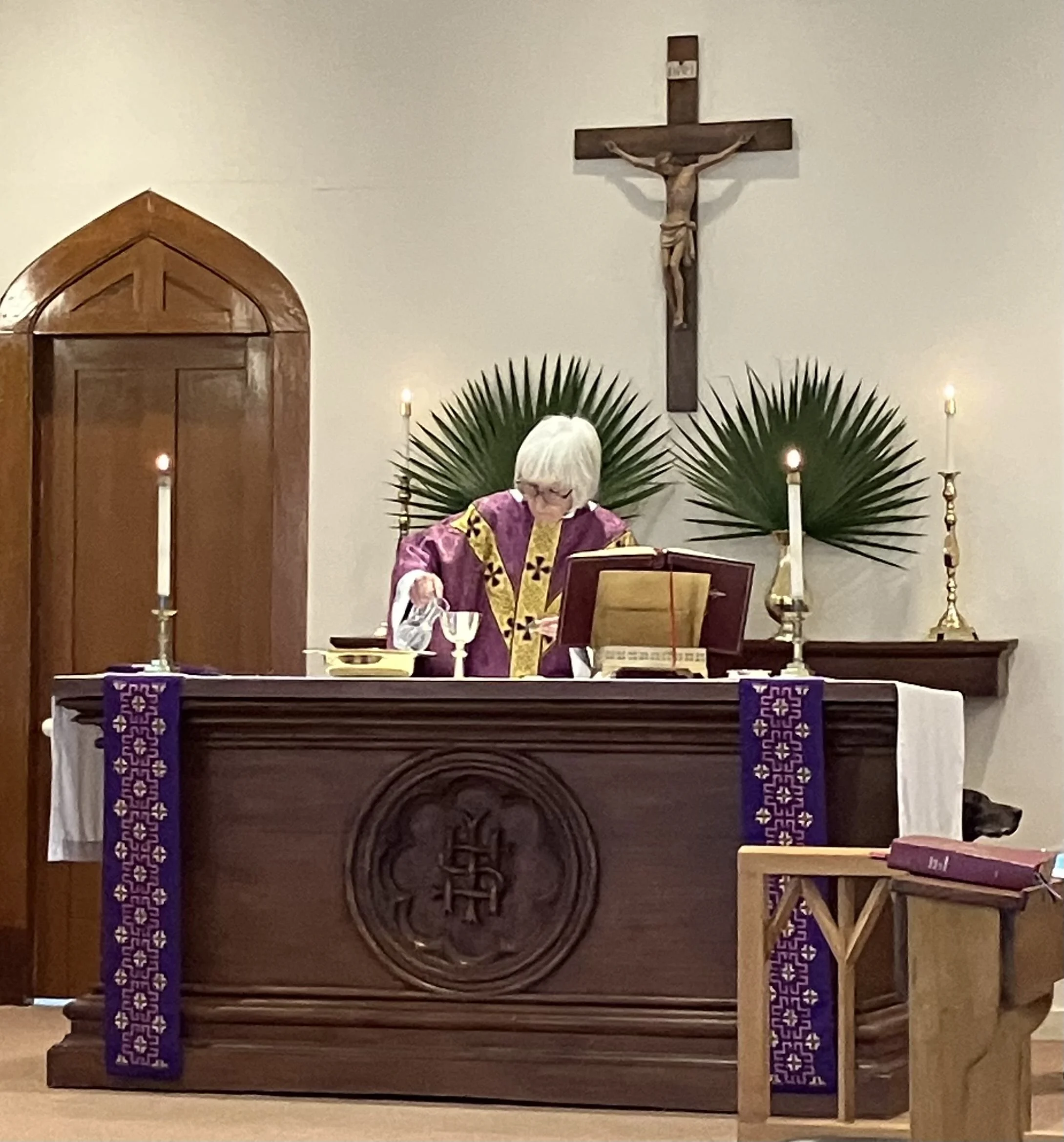 A priest at an altar during a Catholic mass, wearing purple vestments, with candles, a crucifix on the wall, and religious symbols in the background.