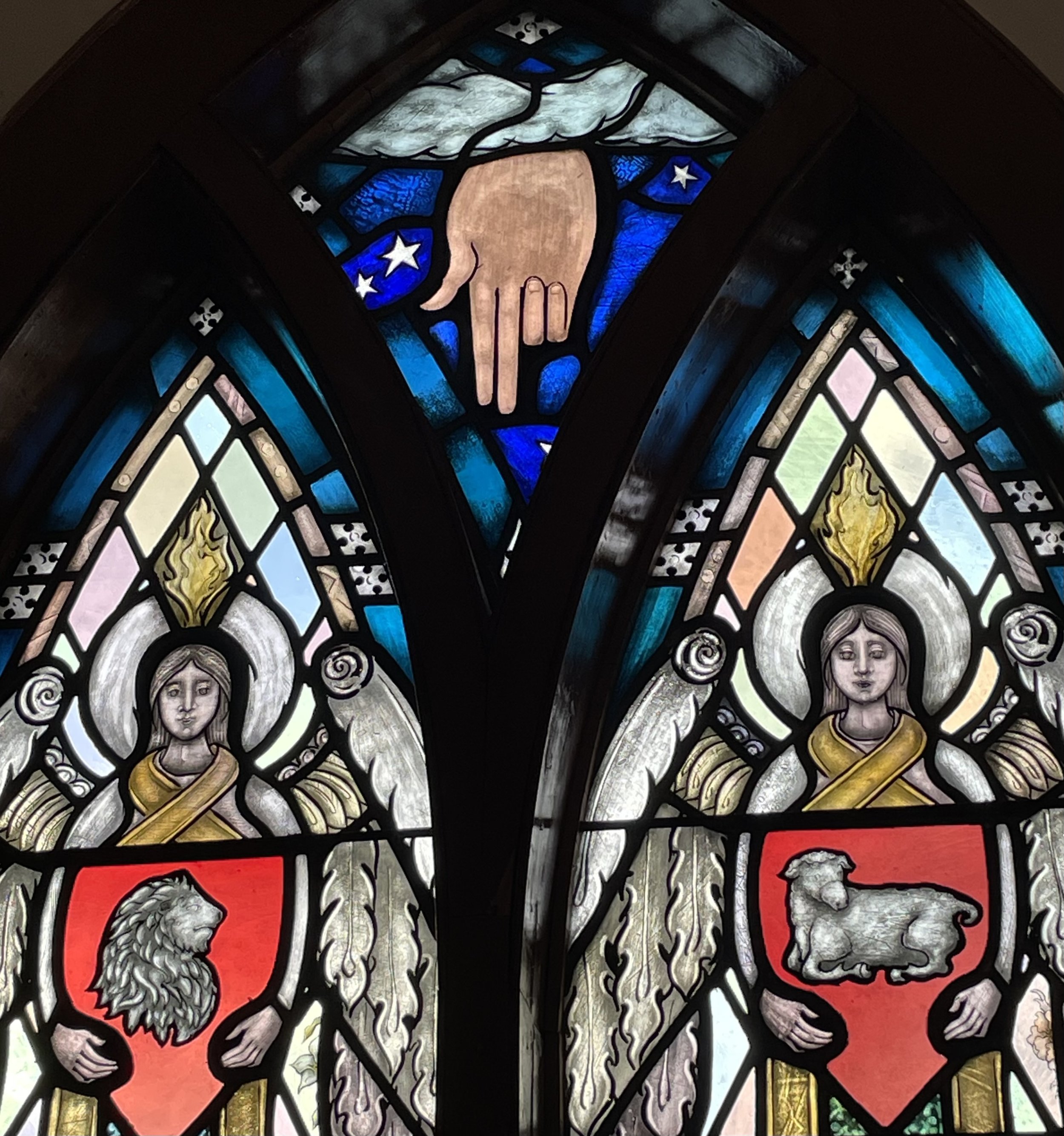 Section of stained glass window with two angels and a hand pointing downward. One angel is holding a shield with a lion and the other a lamb.