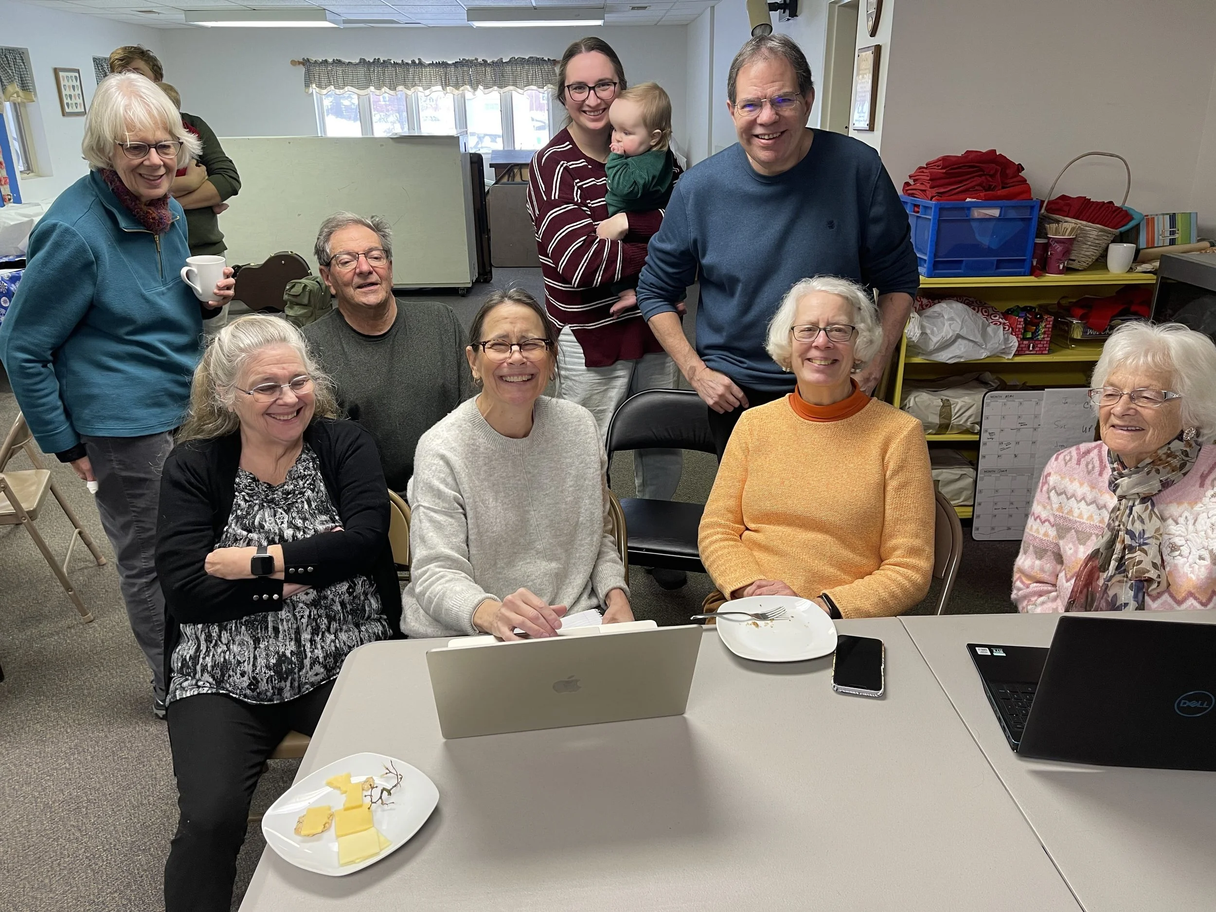 A group of 9 men and women and one baby looking at Mac computer to see new website. Website designer Amy Soutter has biggest smile!