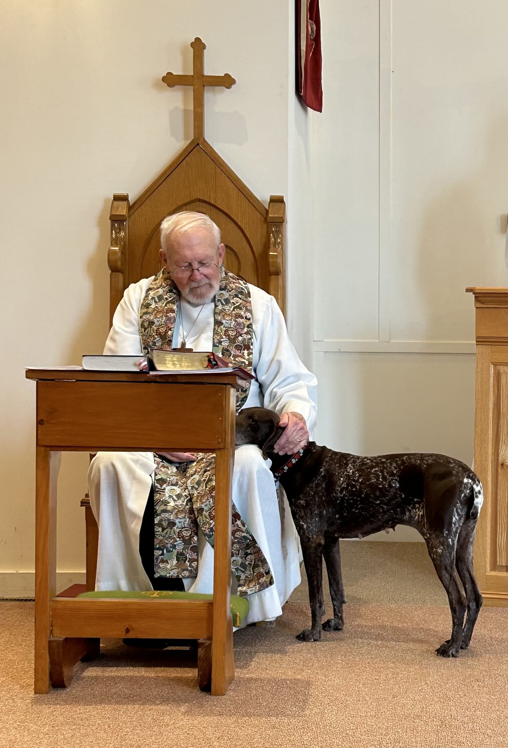 Elderly priest sitting in wooden chair decorated with a carved cross petting a brown dog.