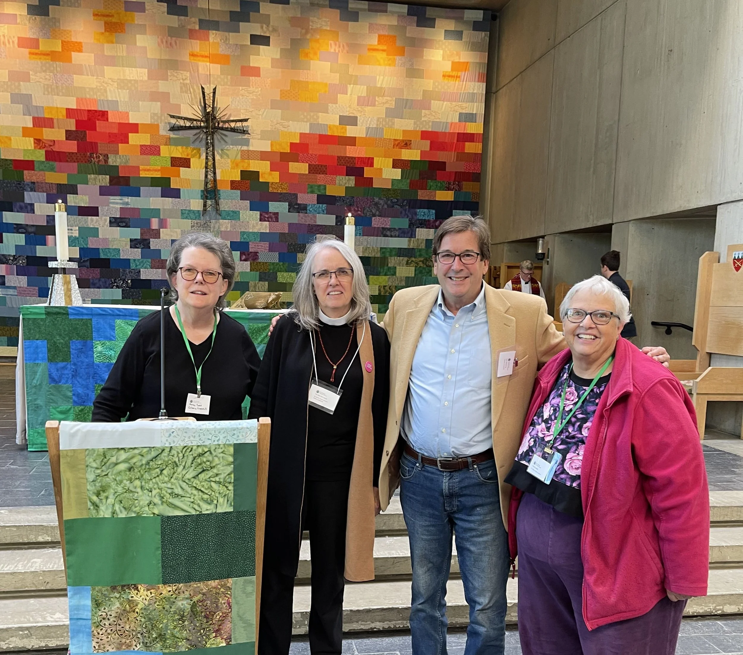 3 Calvary and St. James vestry members (2 women and 1 man) and Rev. Kim Hardy pose after making a presentation at St. Paul’s Cathedral, Burlington. Cross and colorful quilt are in the background.