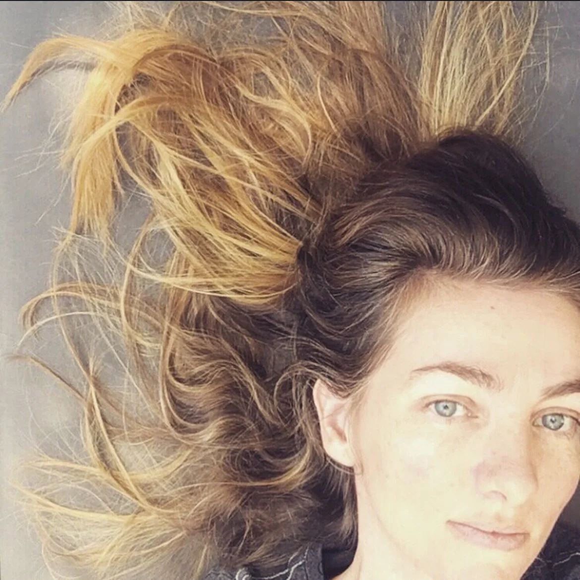 A woman with blue green eyes and light golden skin, lying down with her head turned to the side. She has auburn hair with natural blonde highlights from the sun, spread out around her head.