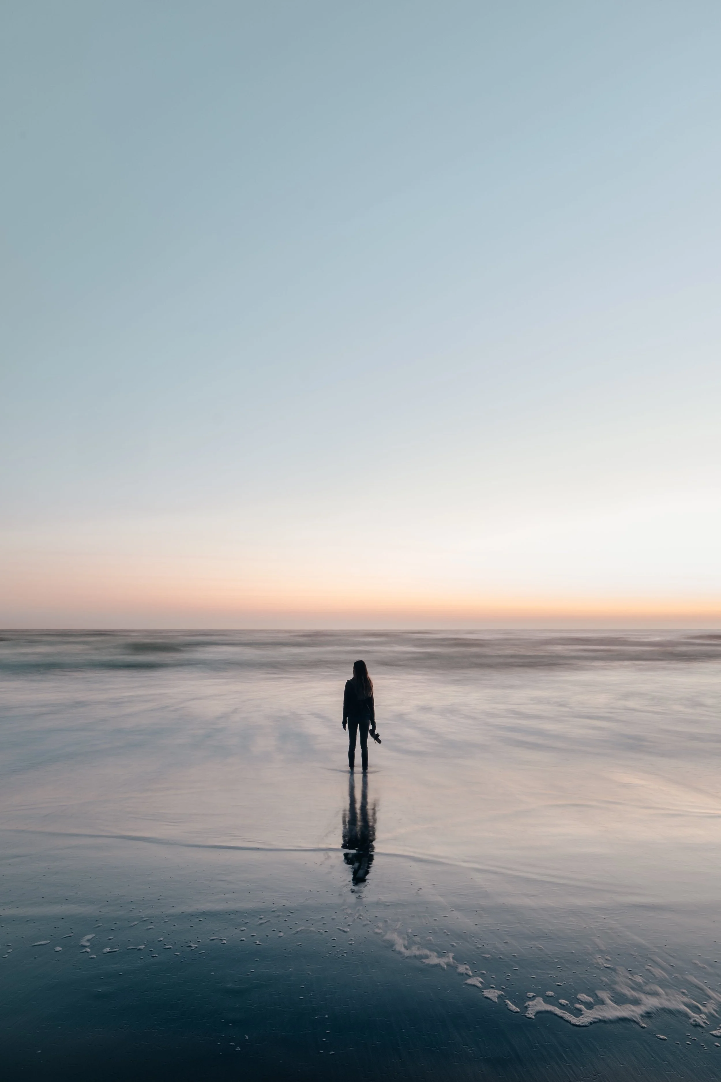 A person walking along a calm, reflective beach at sunset or sunrise, with a pastel-colored sky and gentle waves.