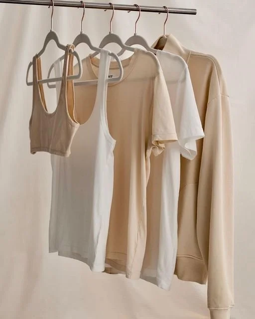 A set of beige and white clothing hanging on a metal rod, including a bra, tank top, t-shirt, and hoodie.