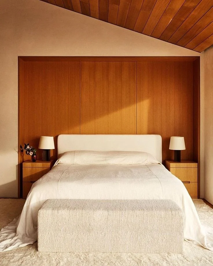A neatly made bed with a white headboard, beige bedding, and a matching foot bench at the foot of the bed. There are two wooden nightstands, each with a white lamp and small decorative items, against a wooden-paneled wall and ceiling.