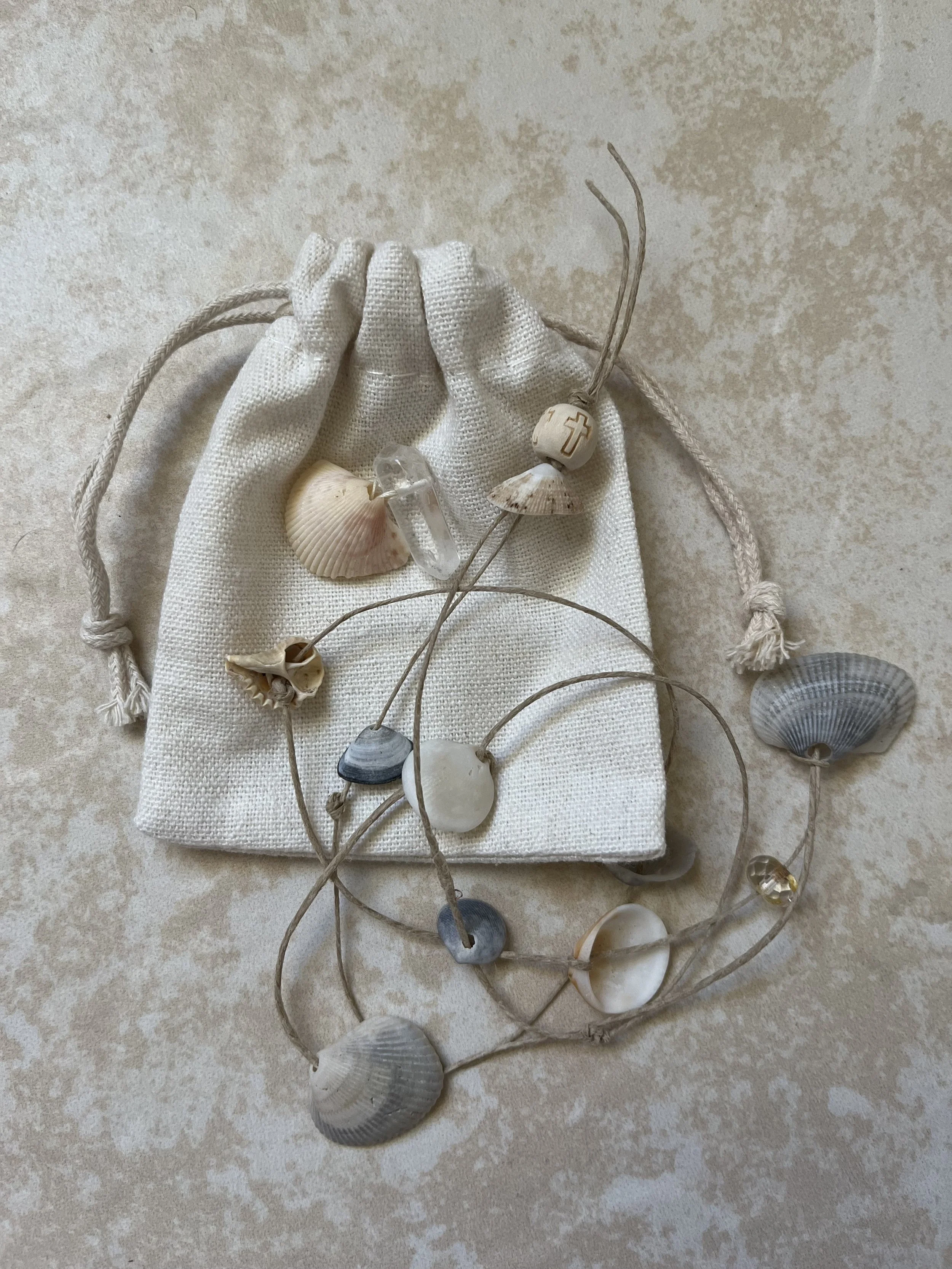 A white fabric drawstring bag on a beige surface, decorated with seashells, a cross bead, and a string of beads with various stones and shells.