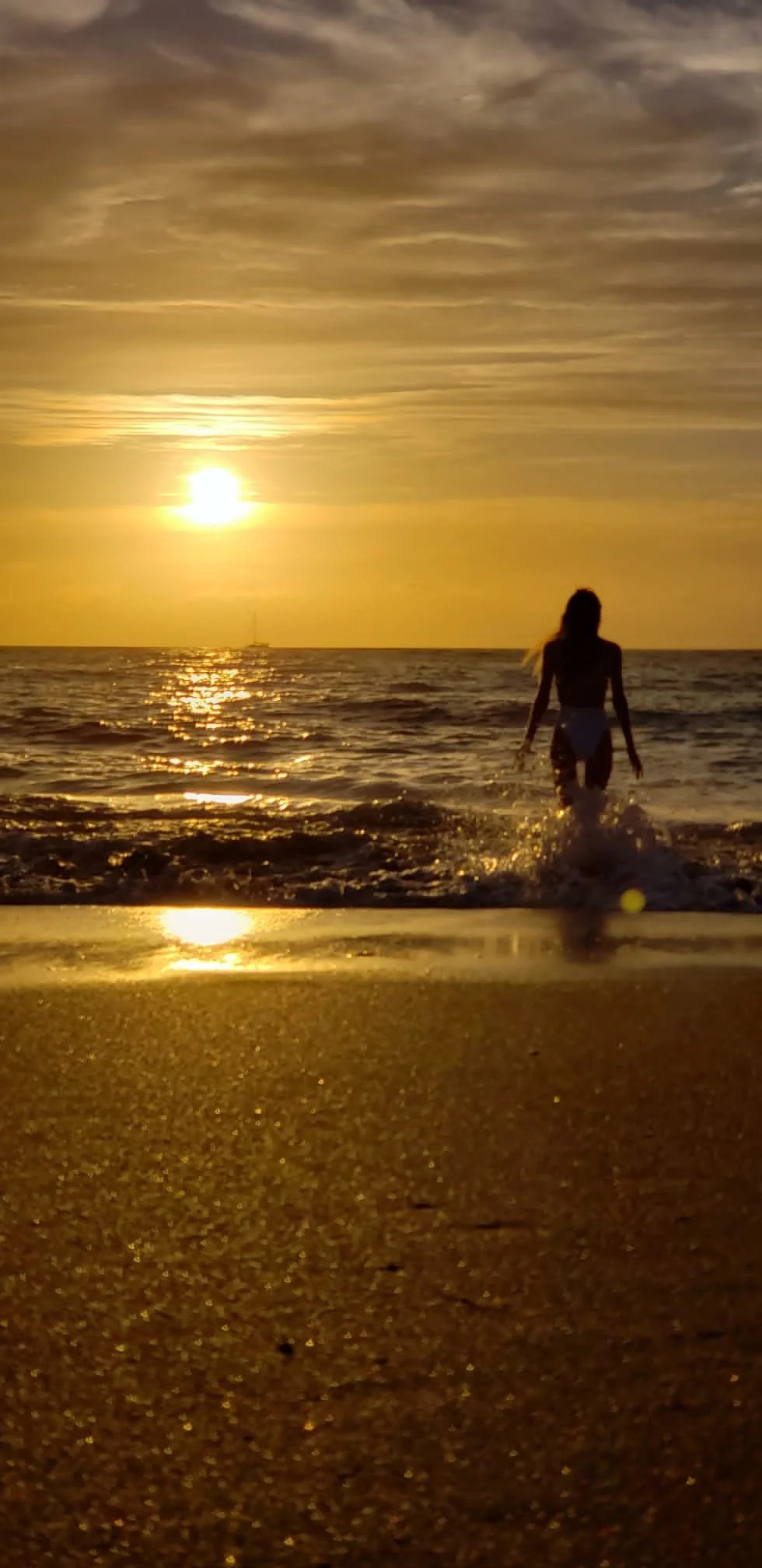A person walking into the ocean at sunrise with golden skies and a sailboat in the distance, capturing a peaceful beach scene.