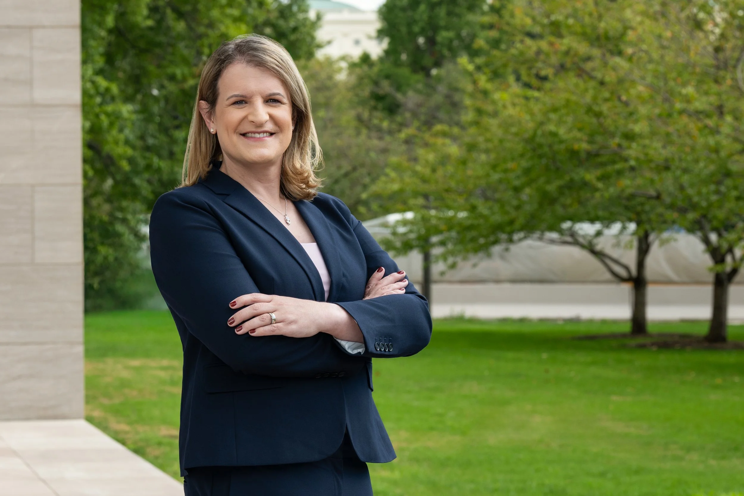 A woman with shoulder-length blonde hair in a navy blue blazer and white shirt, standing outdoors with crossed arms, smiling in front of green trees and a building.