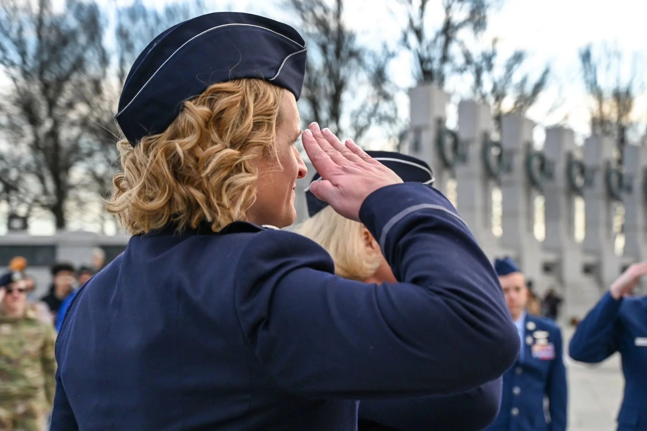 A woman in a navy uniform salutes during a formal event, with several other military personnel in the background. She has curly blonde hair and is saluting with her right hand.