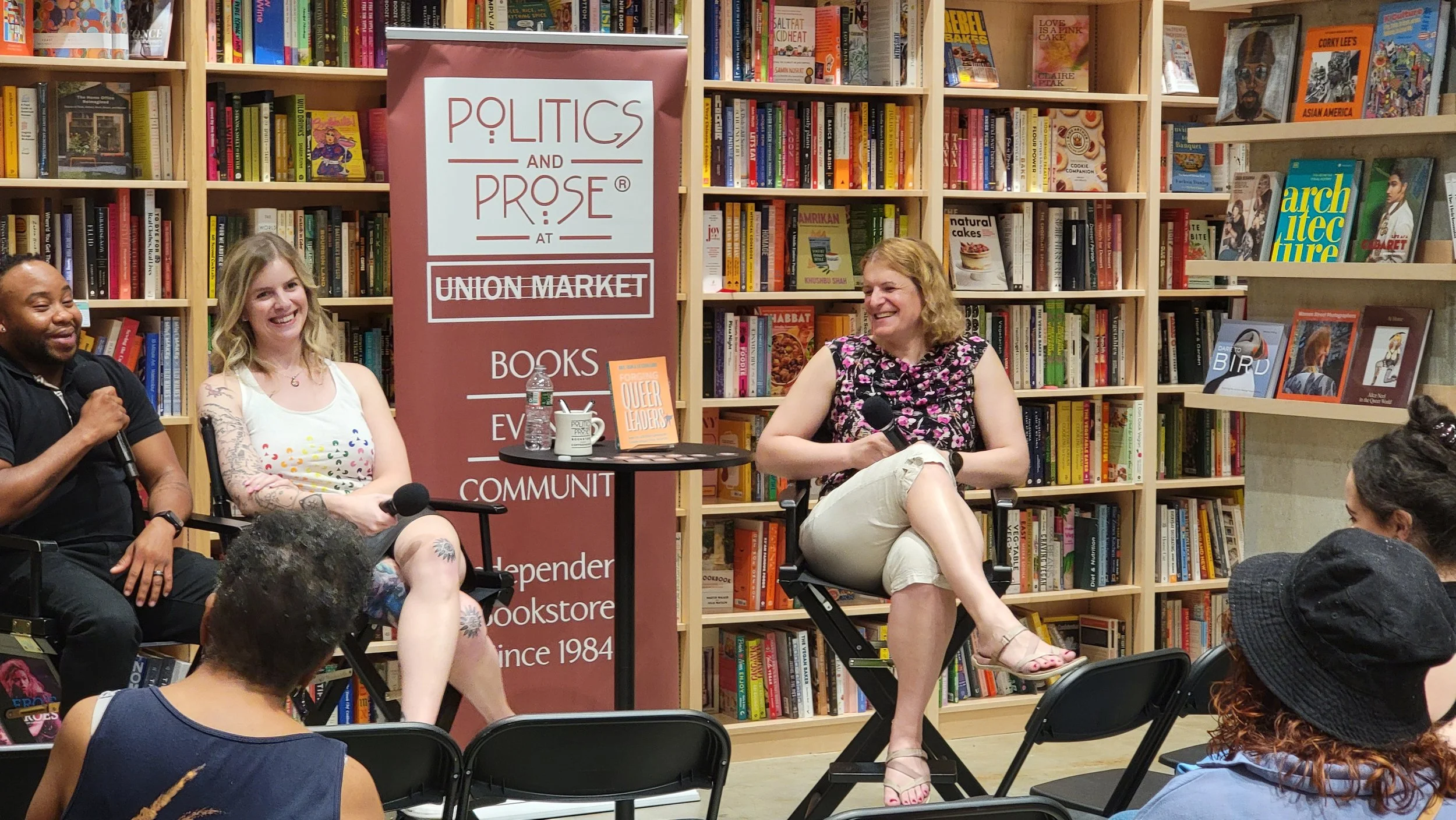 Three women sitting in chairs on stage, holding microphones, and talking in a bookstore or library setting with shelves of books behind them. An audience is seated in front of them.