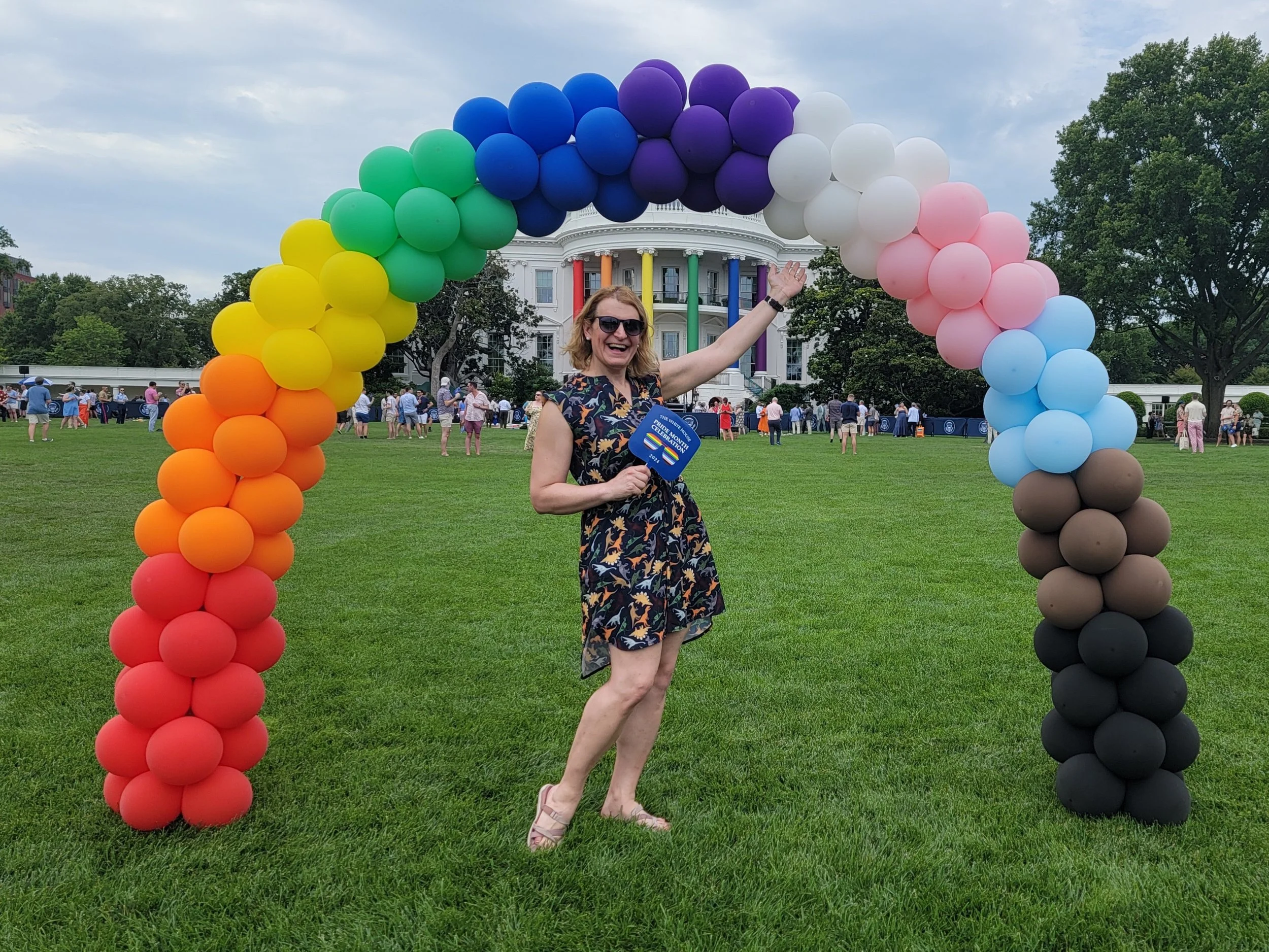 A woman standing on green grass, smiling and pointing upwards, inside a colorful rainbow balloon arch in front of the White House, with a crowd of people in the background.