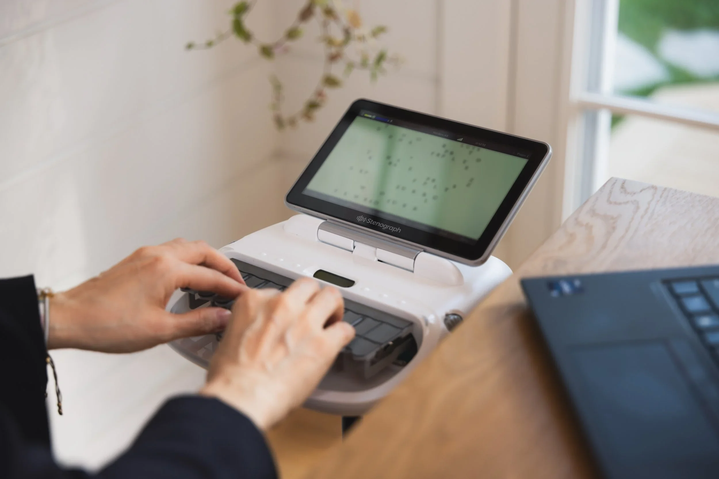 A person typing on a portable braille note-taker device with a built-in refreshable braille display, positioned on a wooden surface near a laptop, with a window and houseplant in the background.