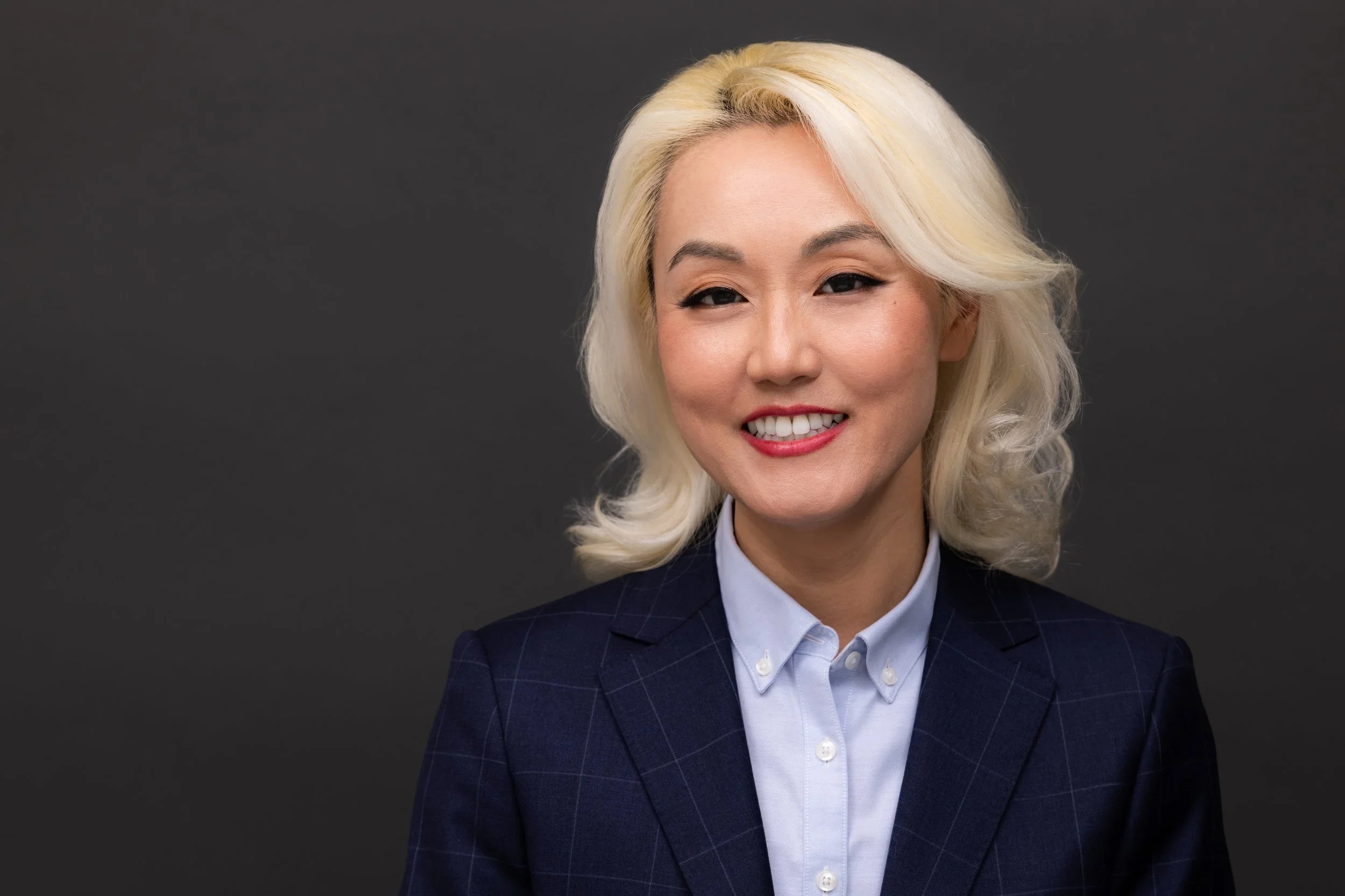 A woman with platinum blonde hair styled in loose waves, wearing a navy blue checked blazer over a light blue button-up shirt, smiling against a dark gray background.