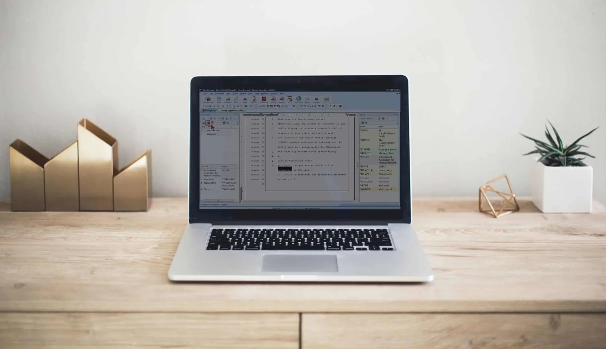 A laptop on a wooden desk displaying a text editing software, with decorative items including gold-colored geometric shapes and a potted plant on either side.