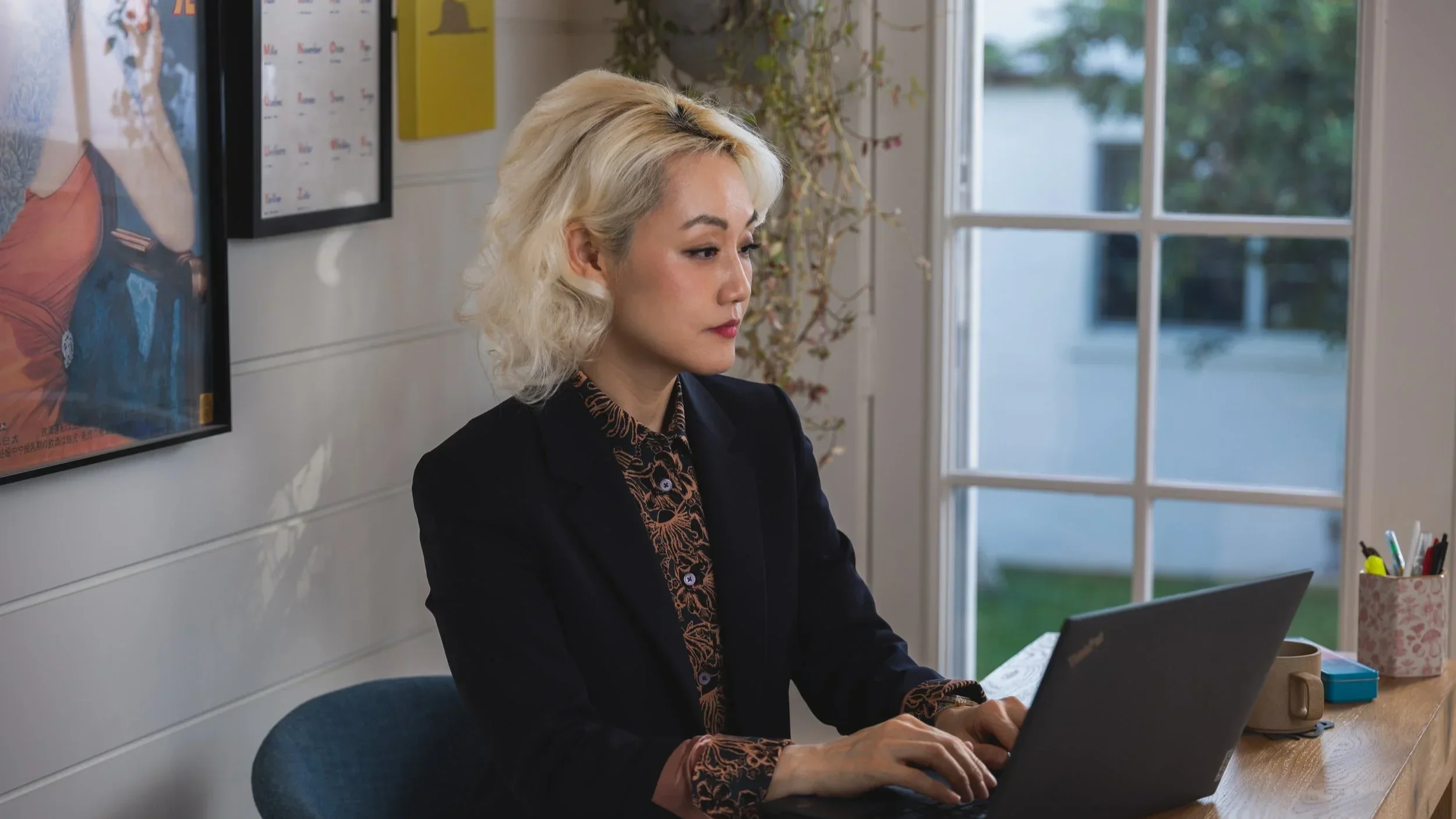 A woman with platinum blonde, curly hair working on a laptop at a desk near a window in a home office.