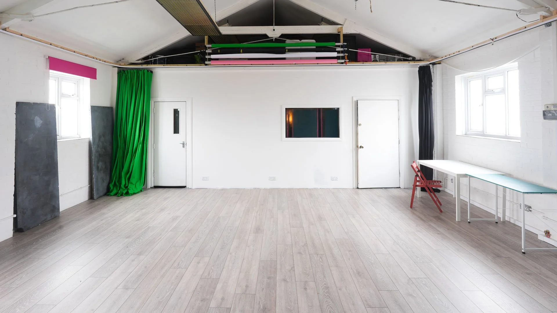 Empty room with white brick walls, large windows, a white table, a red chair, green and black curtains, and studio equipment stored above.