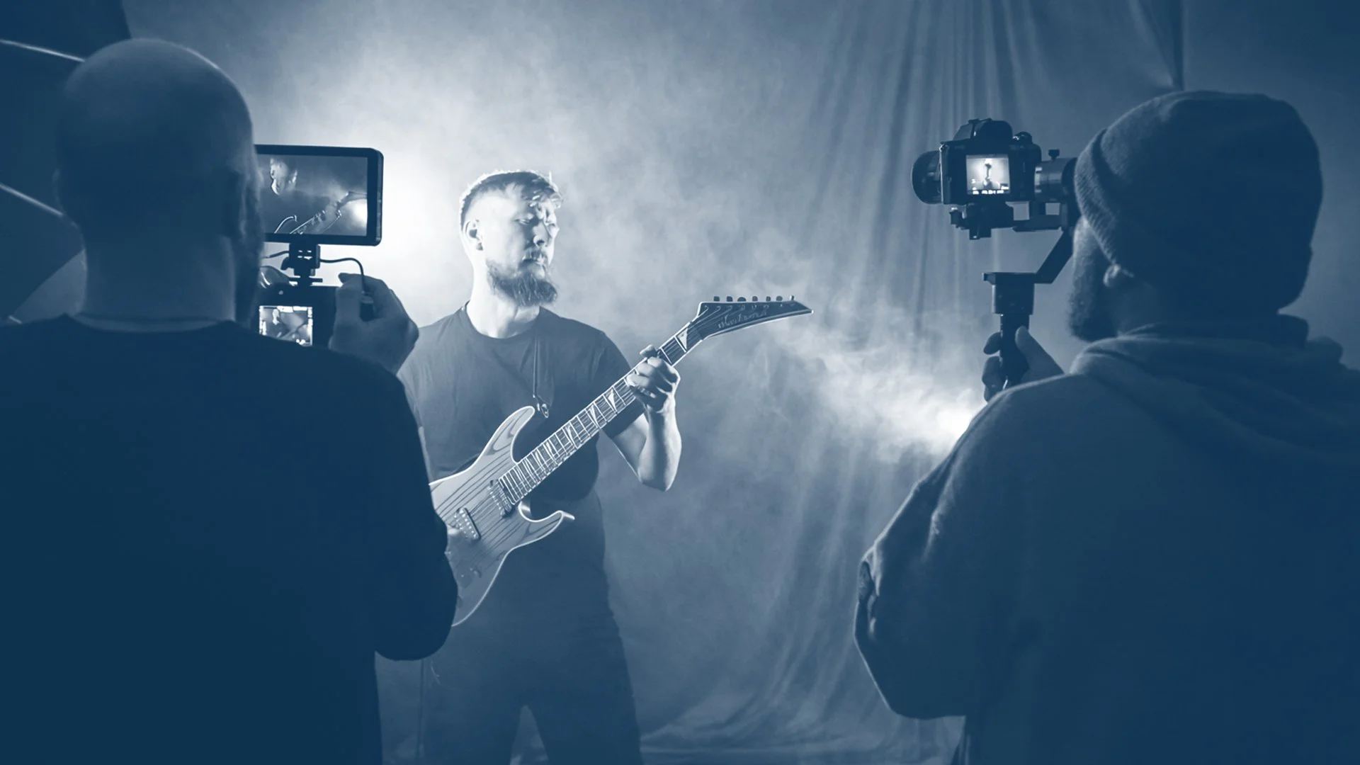 A man with a beard and a guitar playing in a recording studio, surrounded by two videographers filming him with cameras.