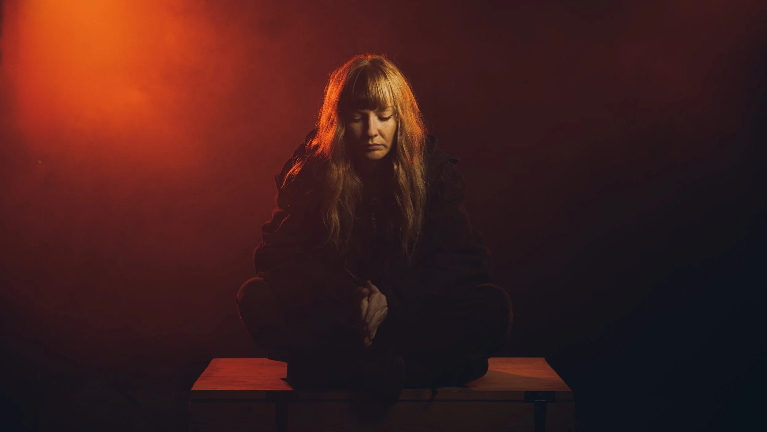 A woman with long blonde hair sits cross-legged on a wooden bench against a dark background, illuminated by a red-orange light.
