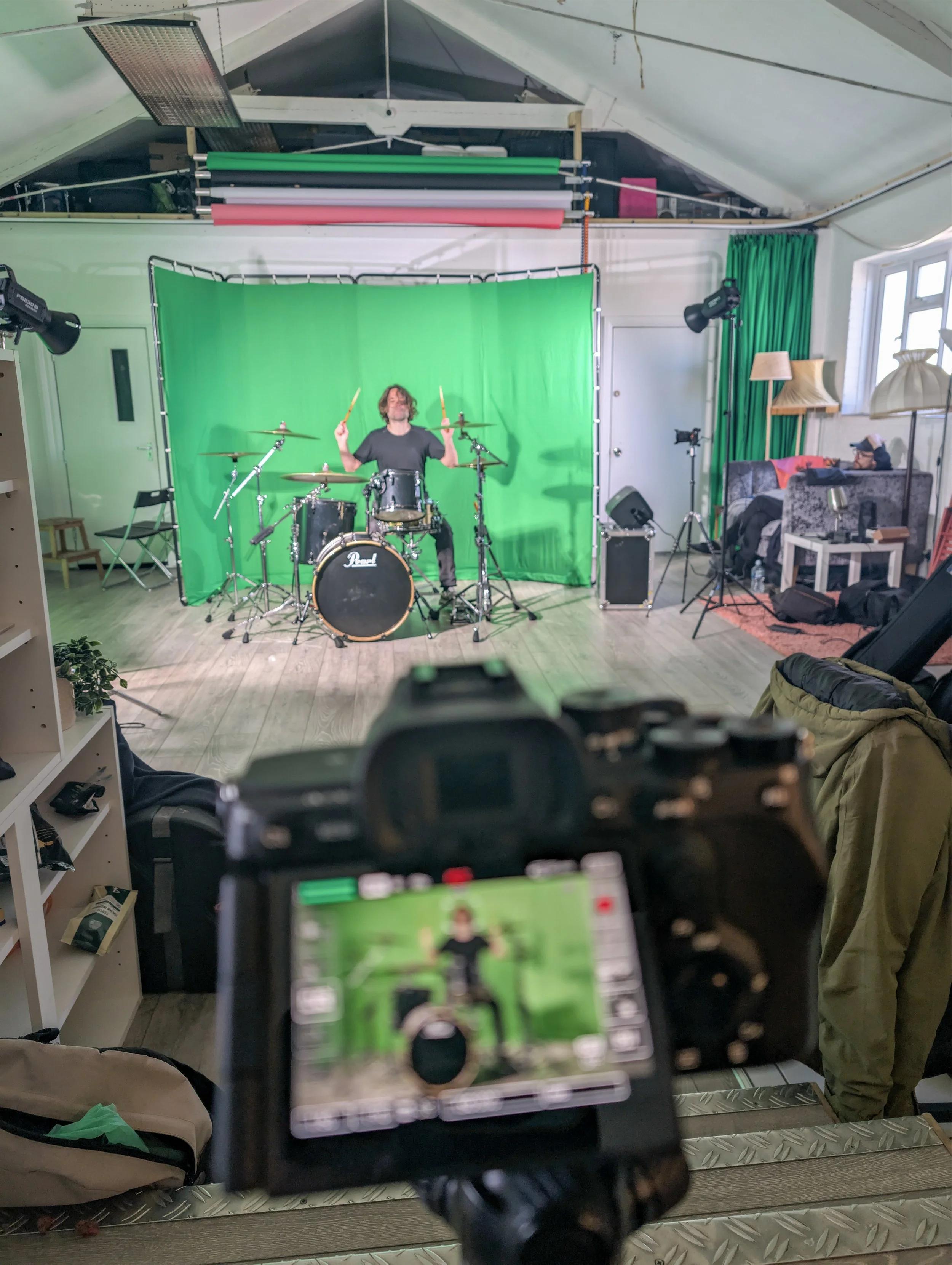 A photo of a recording studio with a green screen backdrop, a drummer playing a Pearl drum set, and a person sitting on a sofa with headphones on. The camera in the foreground is focused on the drummer.