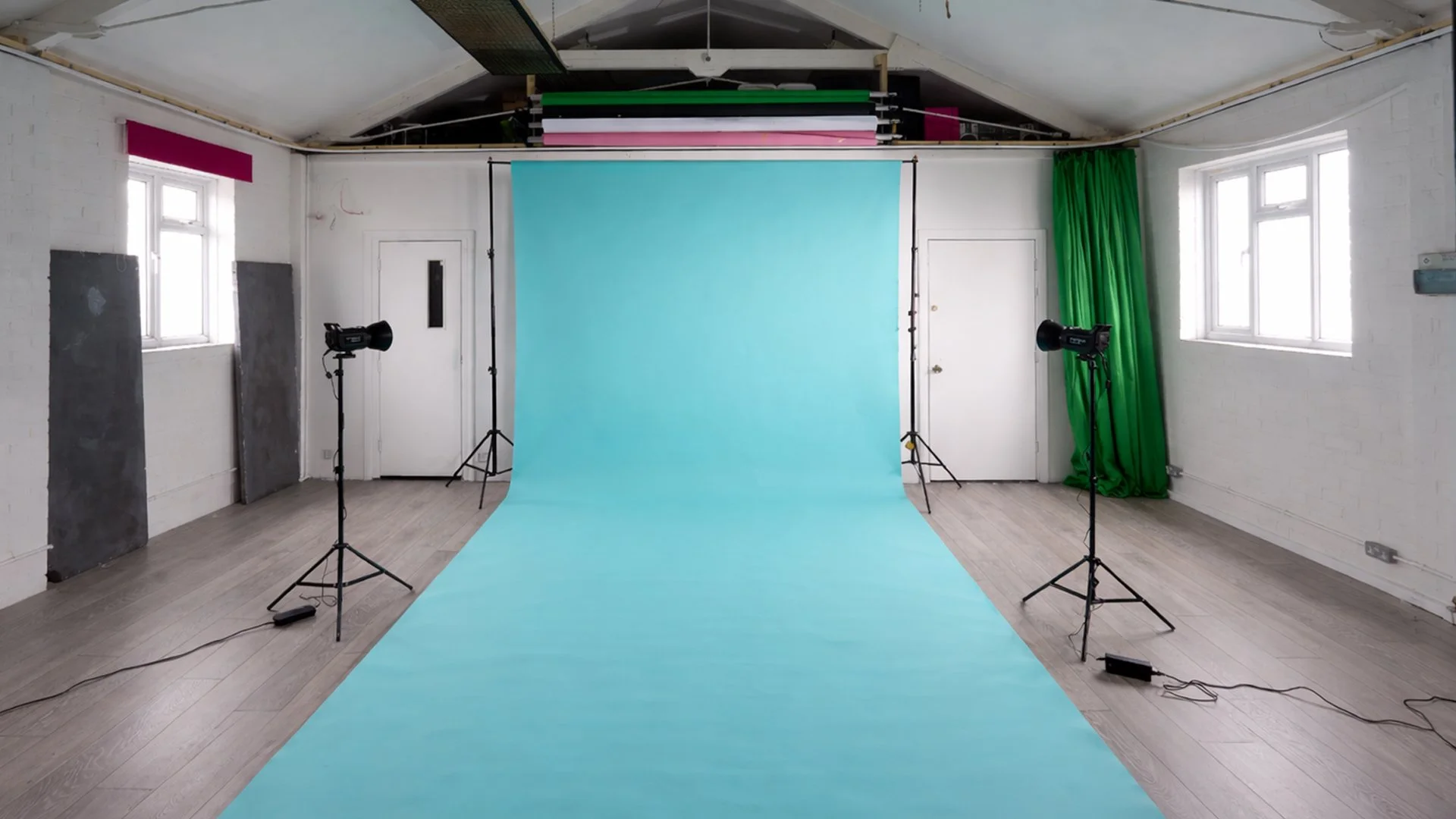 Empty photography studio with light blue backdrop, two studio lights on tripods, green curtain on the right, windows on both sides, and a wooden floor.