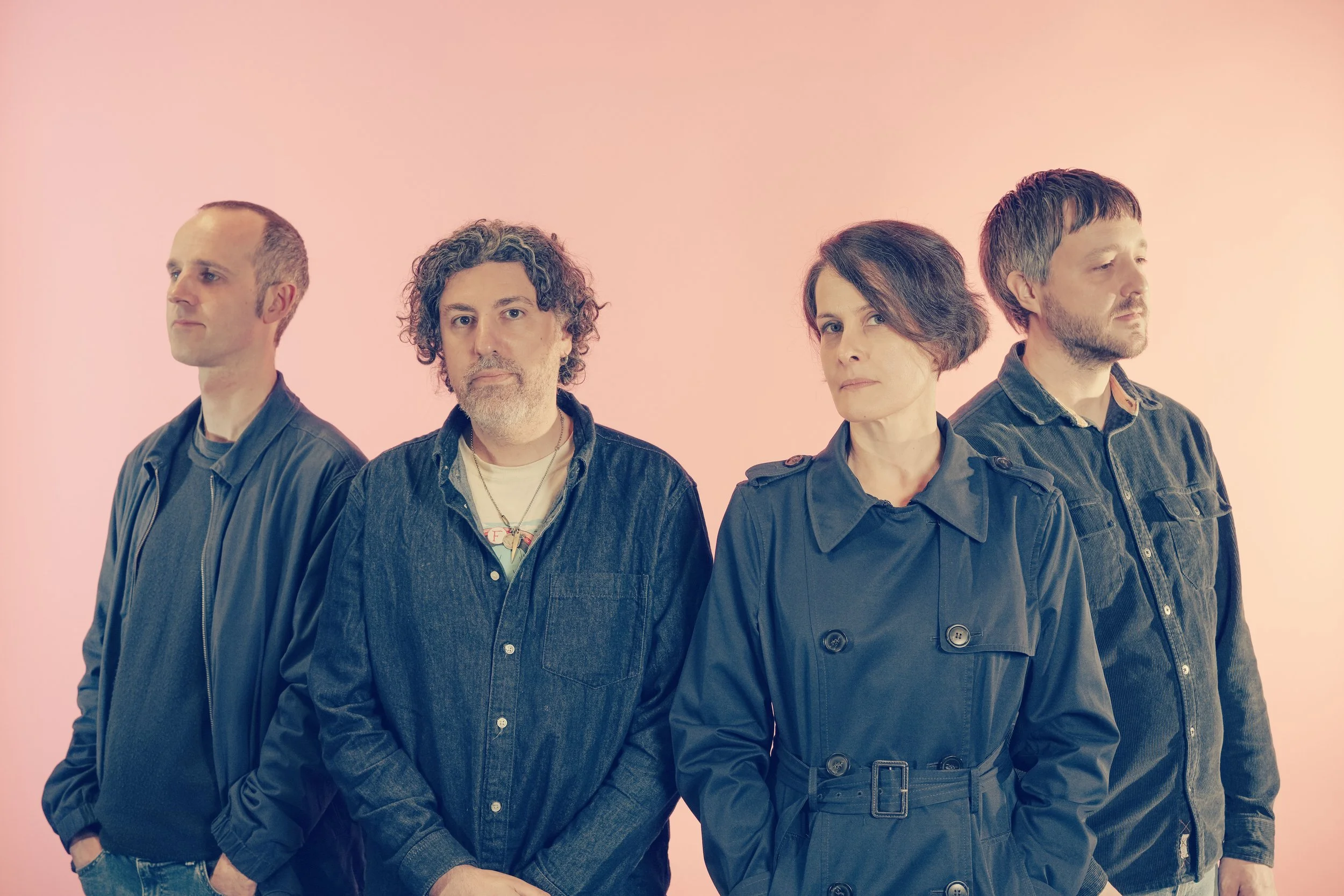 Four people standing in a row against a pink background, facing different directions with serious expressions.