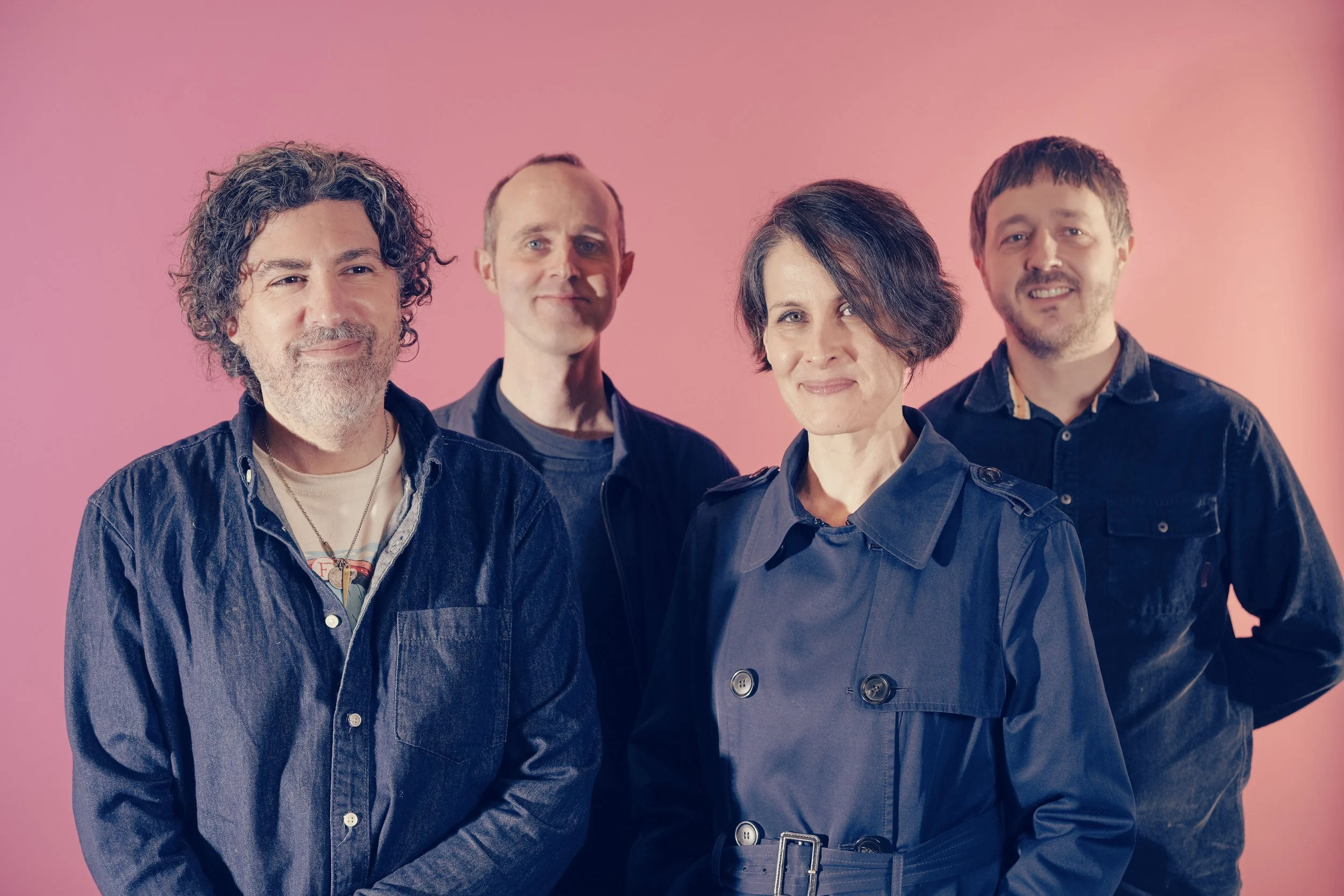 Group of four people standing against a pink background, smiling at the camera.