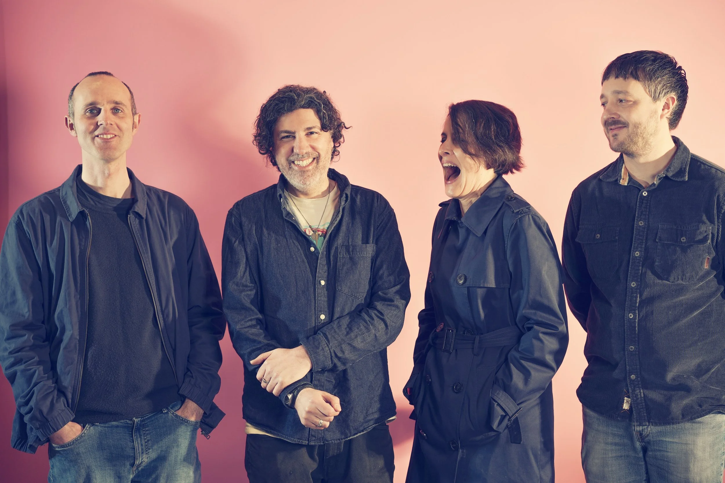 Four people standing against a pink background, laughing and smiling, dressed casually.