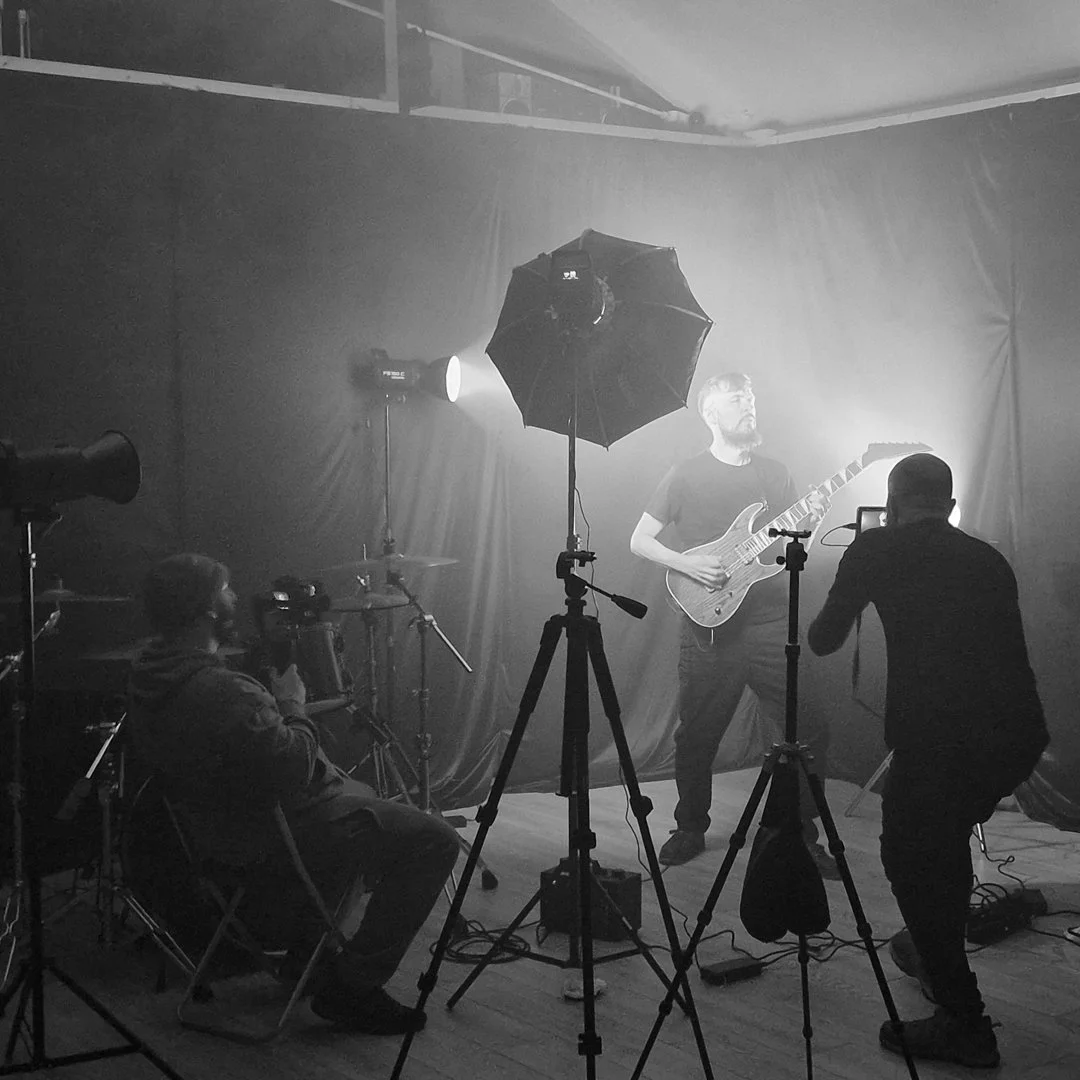 A black and white photo of a music video or photo shoot with a man playing an electric guitar on a dark stage with lighting equipment, cameras, and crew members around him.