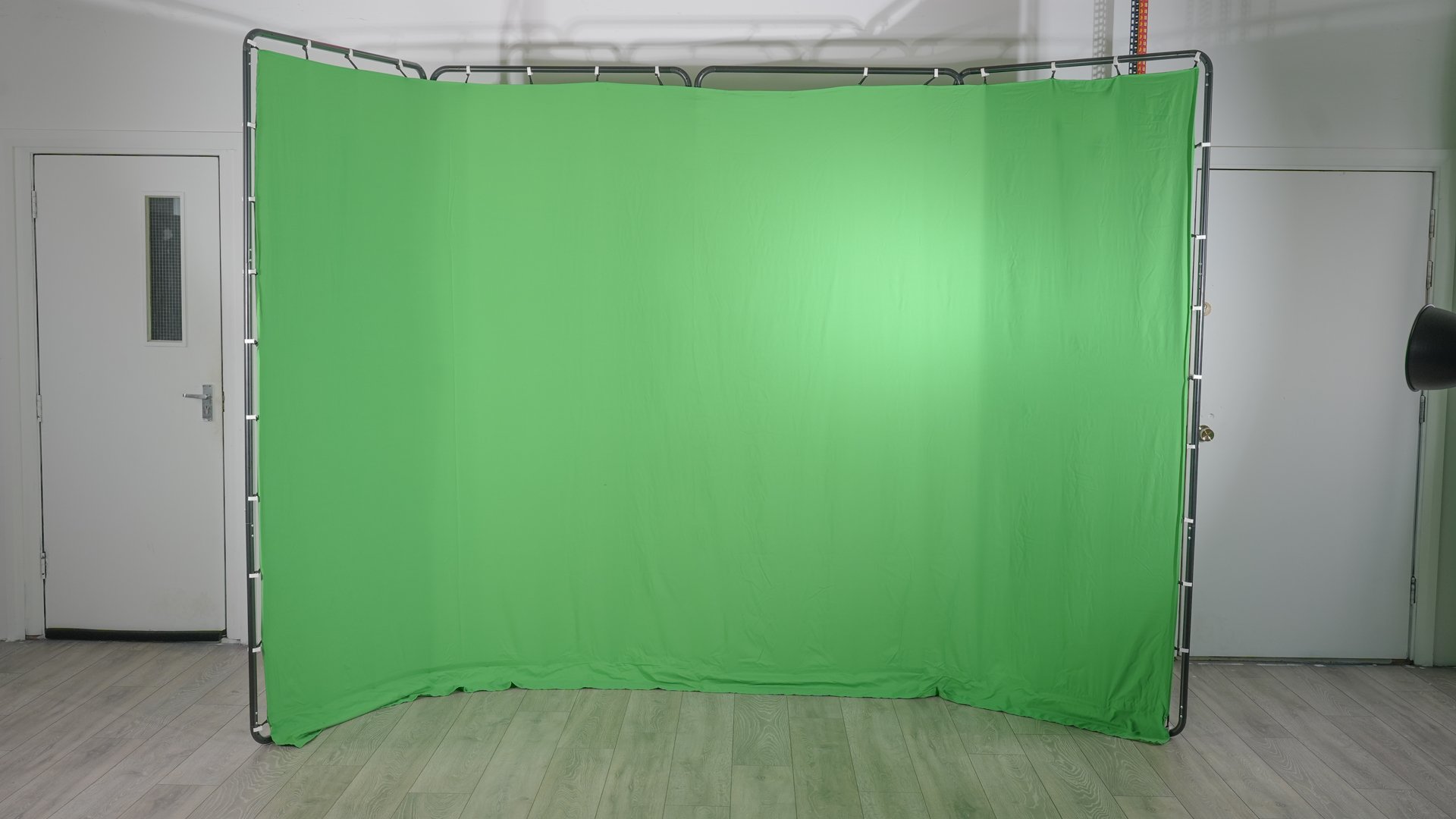 Indoor photo of a green screen backdrop on a metal frame, positioned on a light-colored wood floor against a plain wall with two doors.