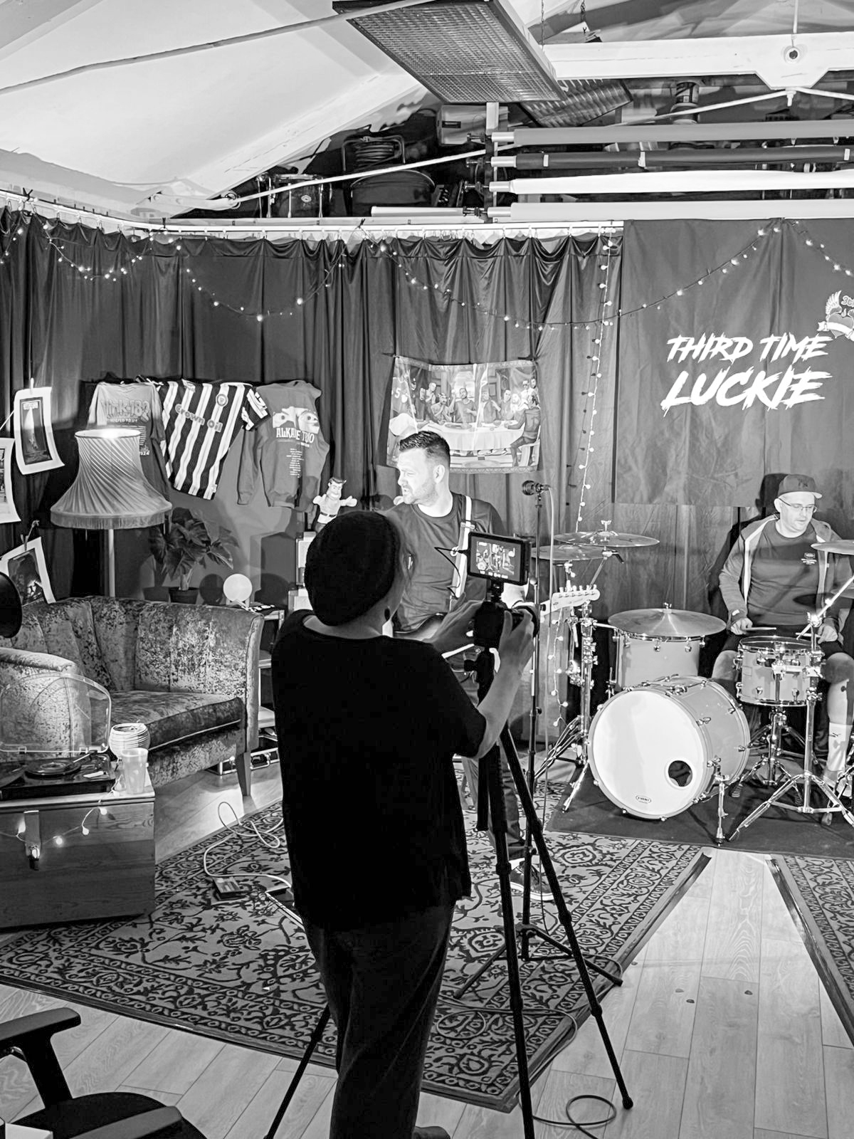 The image shows a black-and-white scene of a recording or filming setup in a cozy studio with a black curtain backdrop. A cameraman is filming two men who are seated behind a drum set, while a third person is filming them with a camera on a tripod. T