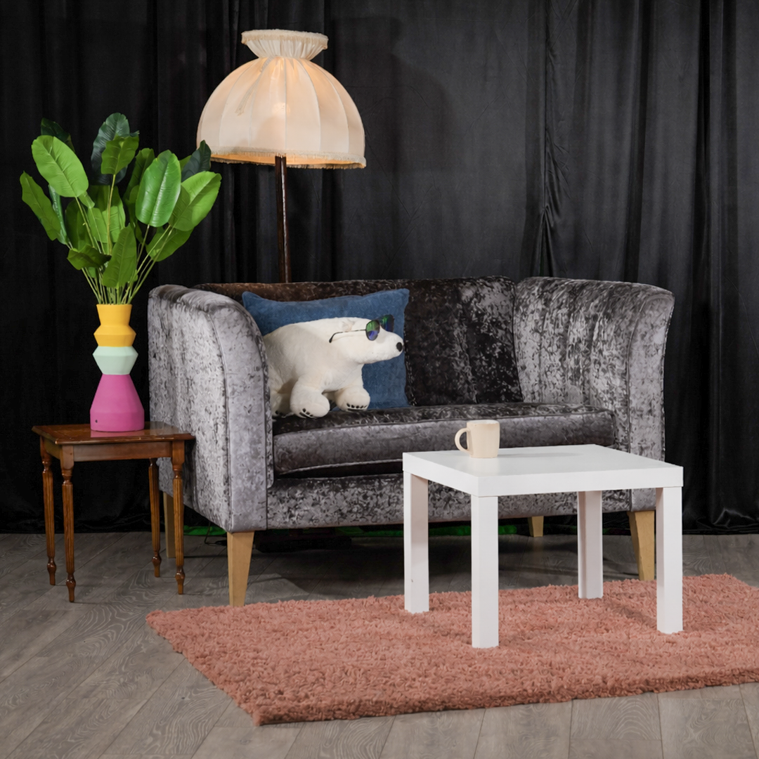 A cozy living room setup with a black velvet couch, a plush polar bear stuffed animal wearing sunglasses, a small white coffee table with a mug, a side table with a colorful geometric vase with green leaves, a floor lamp with a beige shade, and a pin