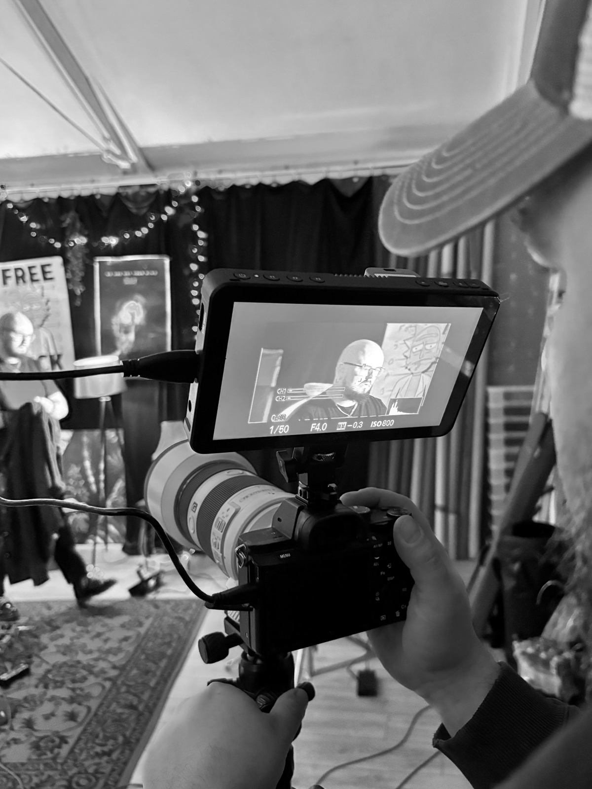 A person filming a theatrical performance with a professional camera on a tripod in a room with dark curtains, banners, and decorative lights.