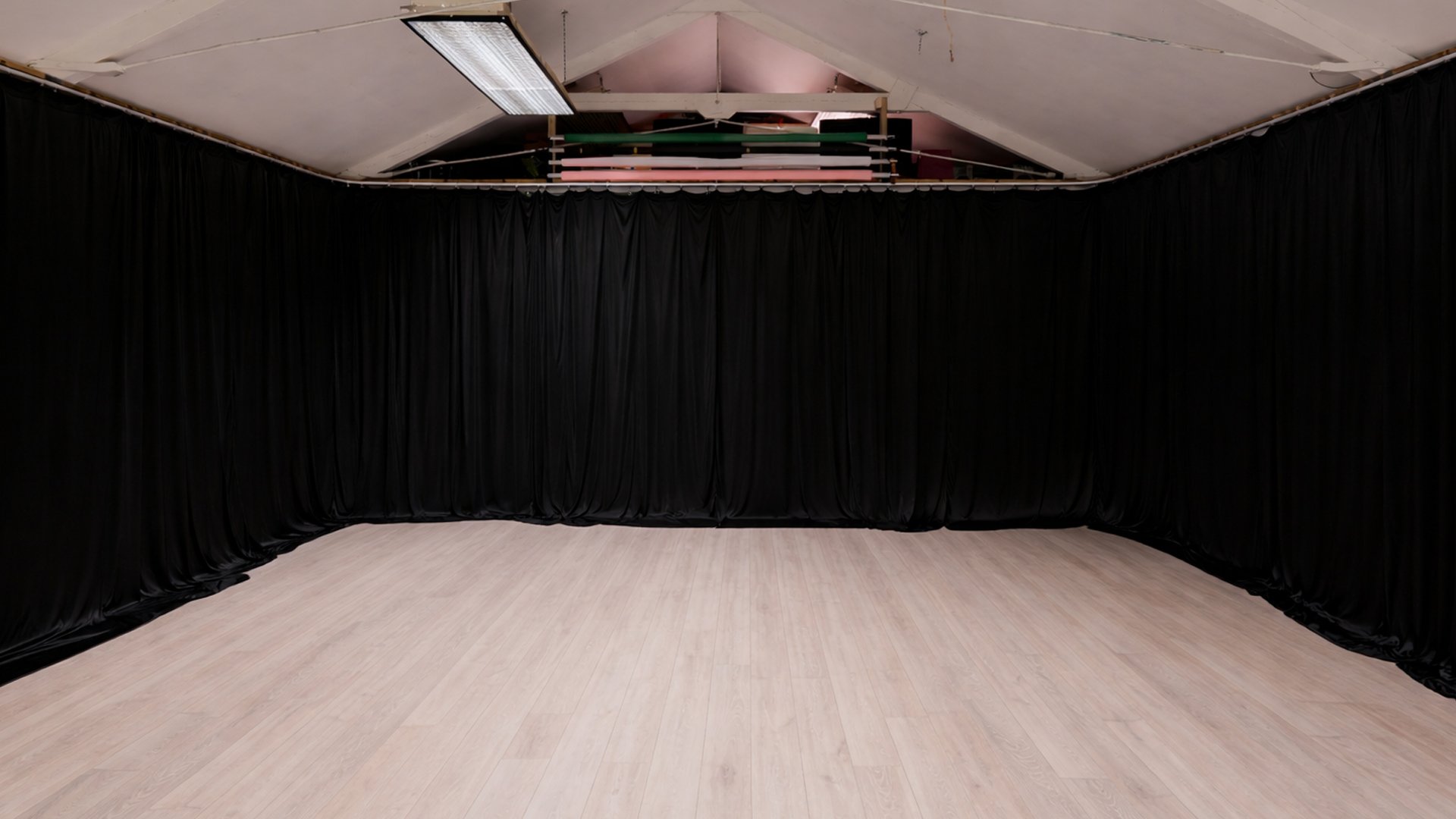 Empty stage with light wood flooring, surrounded by black curtains, with stage equipment stored above.