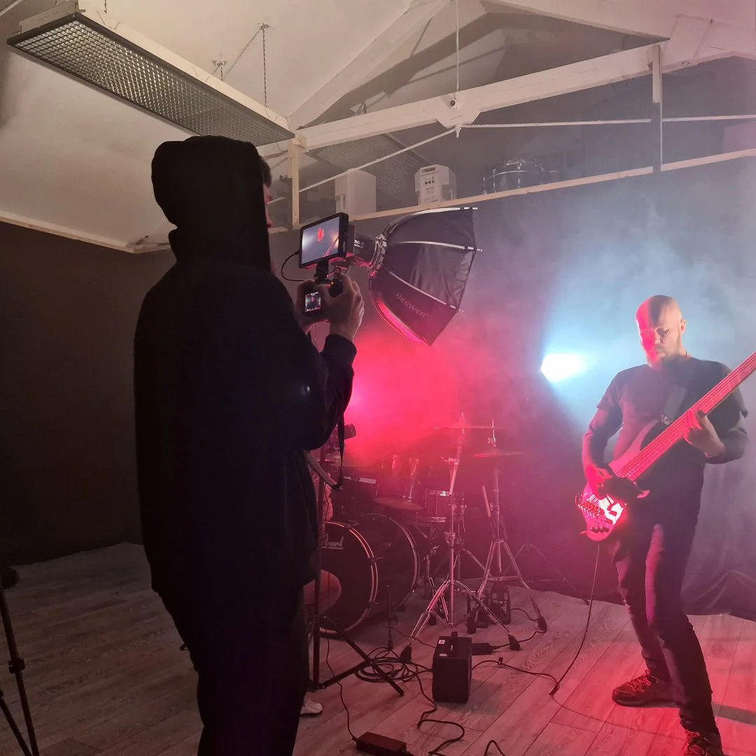 A person in a black hoodie filming a guitarist playing an electric guitar in a music studio with stage lights and smoke.
