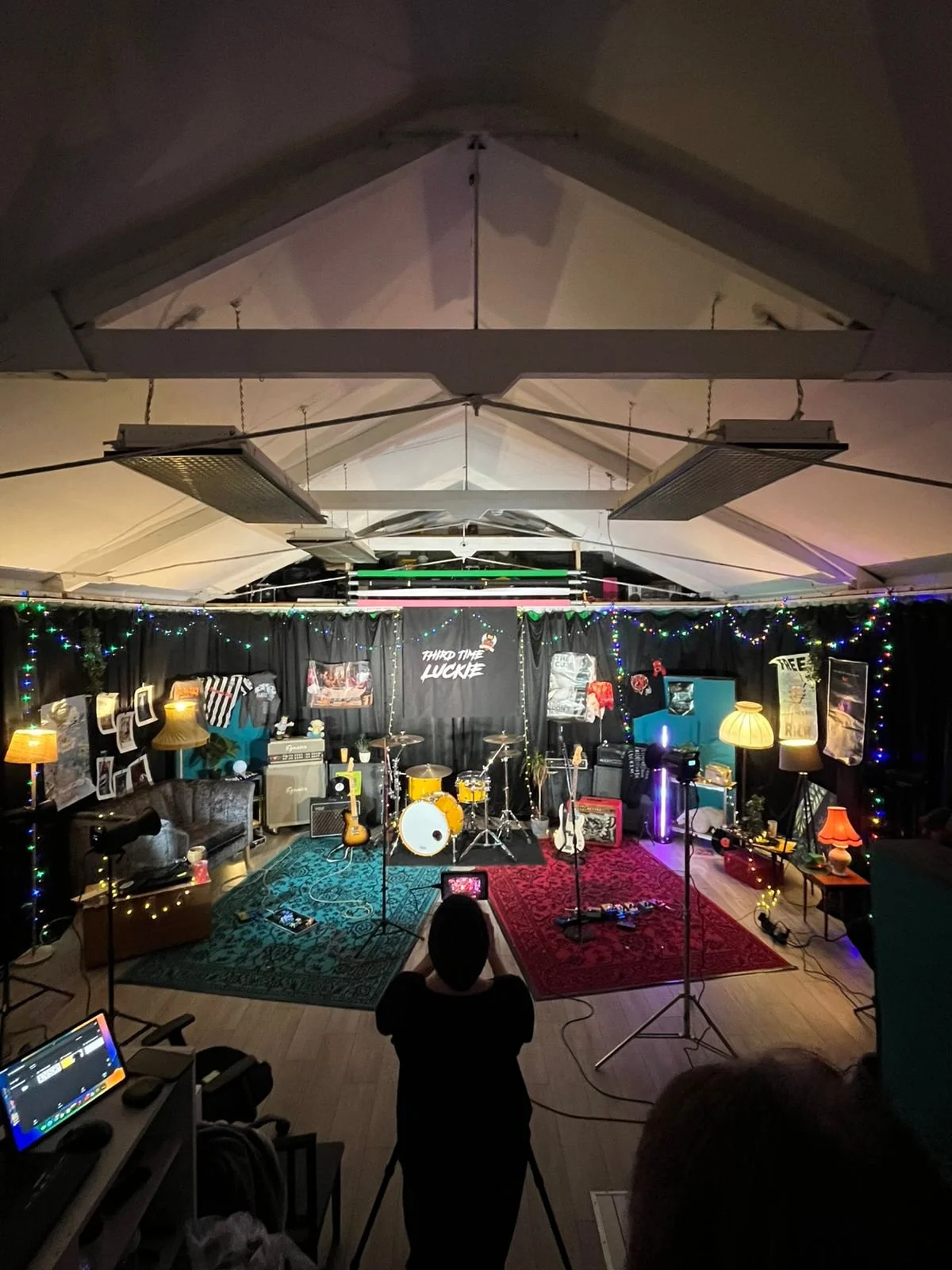 Interior view of a music stage setup with drums, guitars, and amplifiers, surrounded by colorful string lights, lamps, and posters, with a person taking a photo in the foreground.