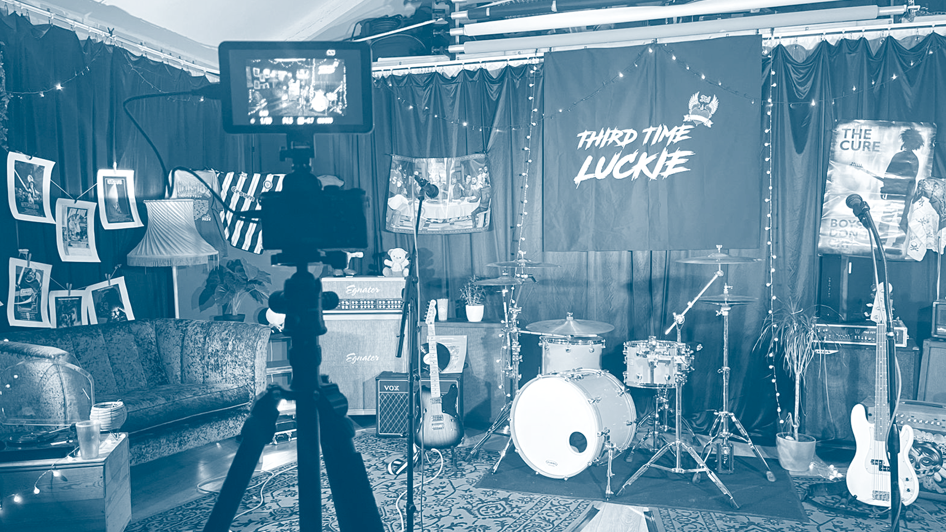 A music studio stage with drums, guitars, microphones, and a backdrop that reads 'Third Time Luckie,' decorated with posters and photographs.