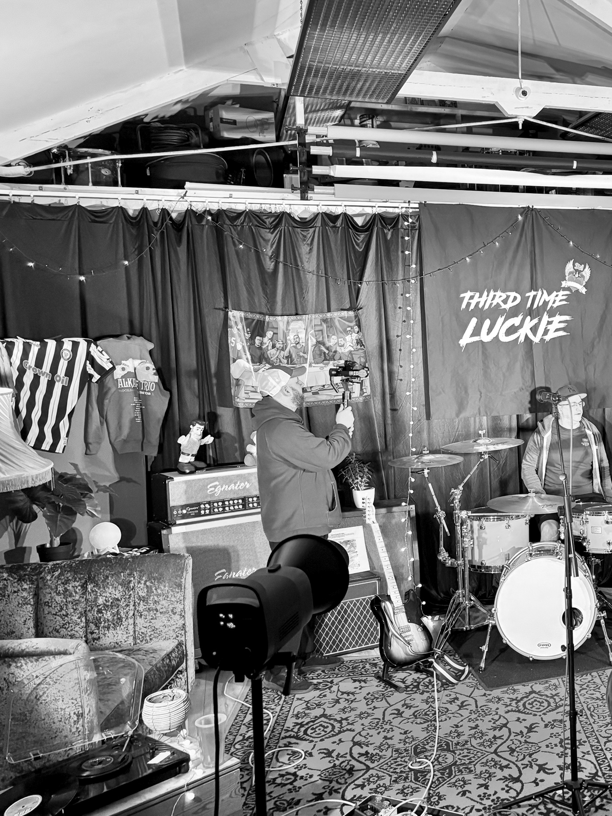 Black and white photo of a music performance stage with a person singing into a microphone, drums, guitar, and various decorations, including a banner that reads 'Third Time Luckie' and hanging lights.
