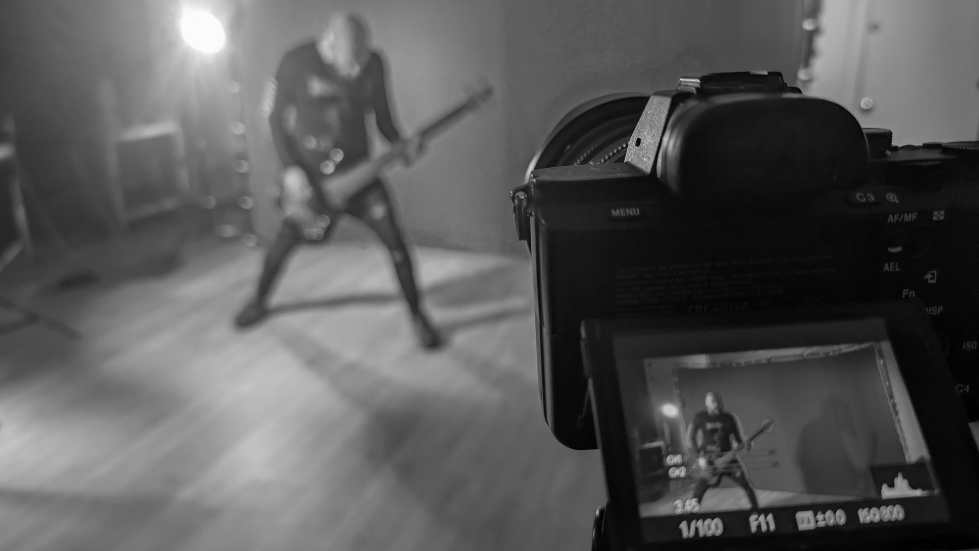 A photographer's camera captures a black and white image of a person playing an electric guitar, posed with legs apart and head down, on a wood floor with a plain wall in the background.