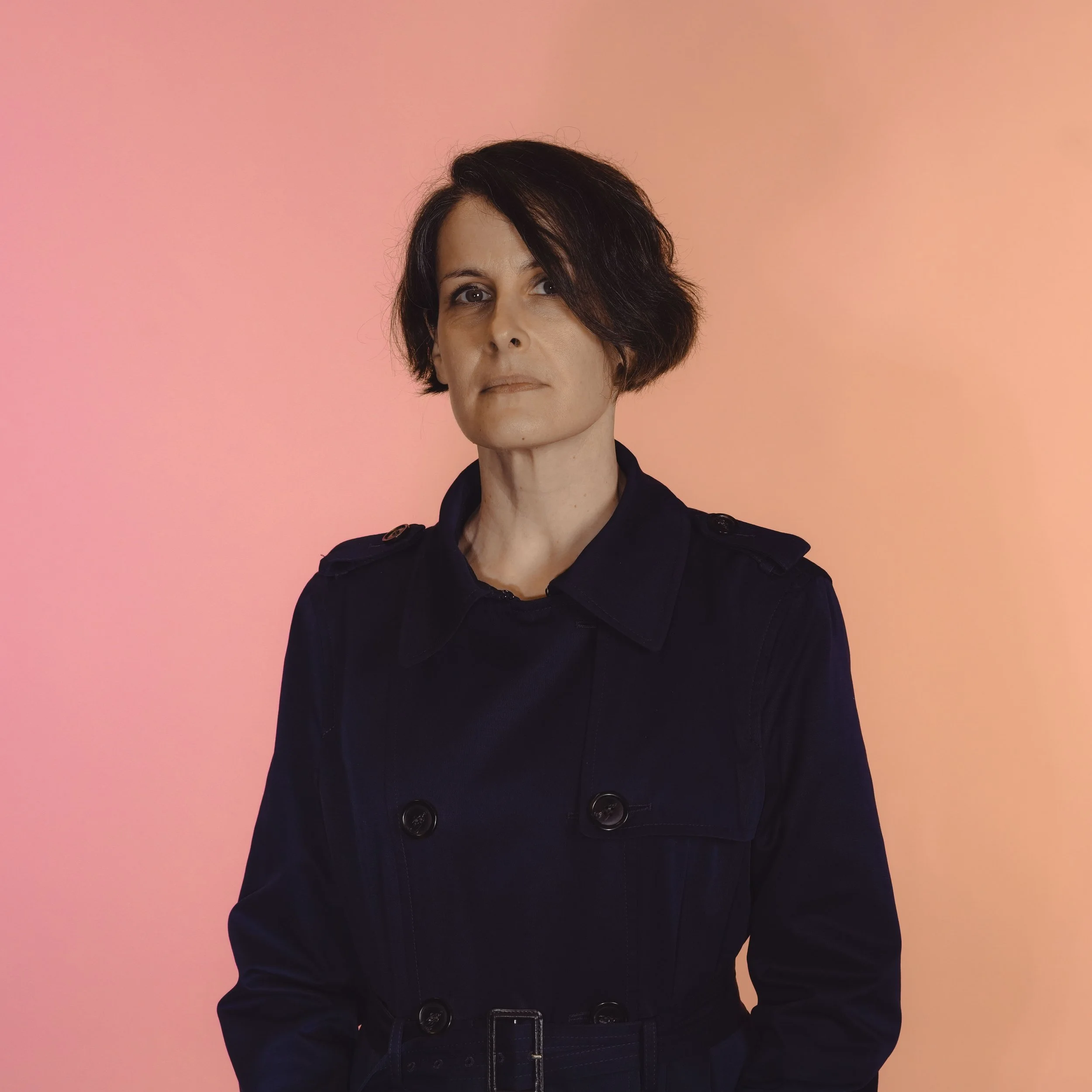 A woman with short dark hair styled in a bob, wearing a black trench coat, standing against a pink and peach gradient background.