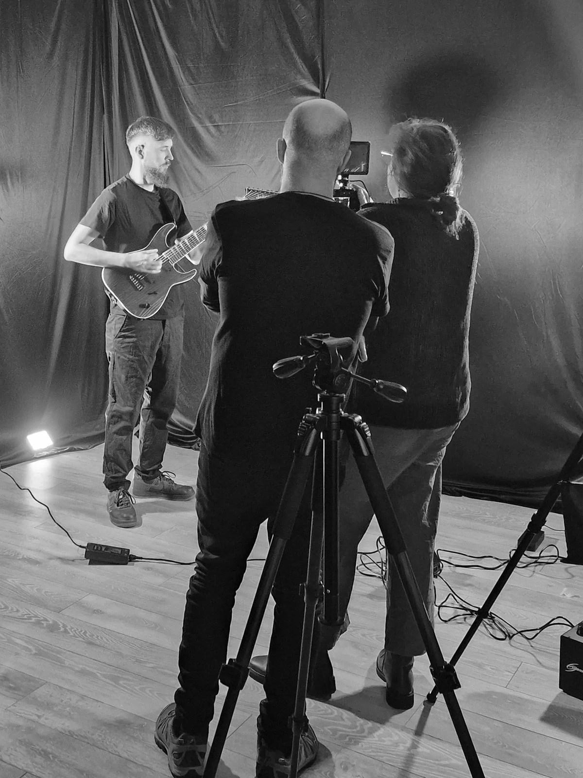 A man plays an electric guitar during a recording session with two women filming him, one with a camera and another with a phone, in a studio with black curtains.