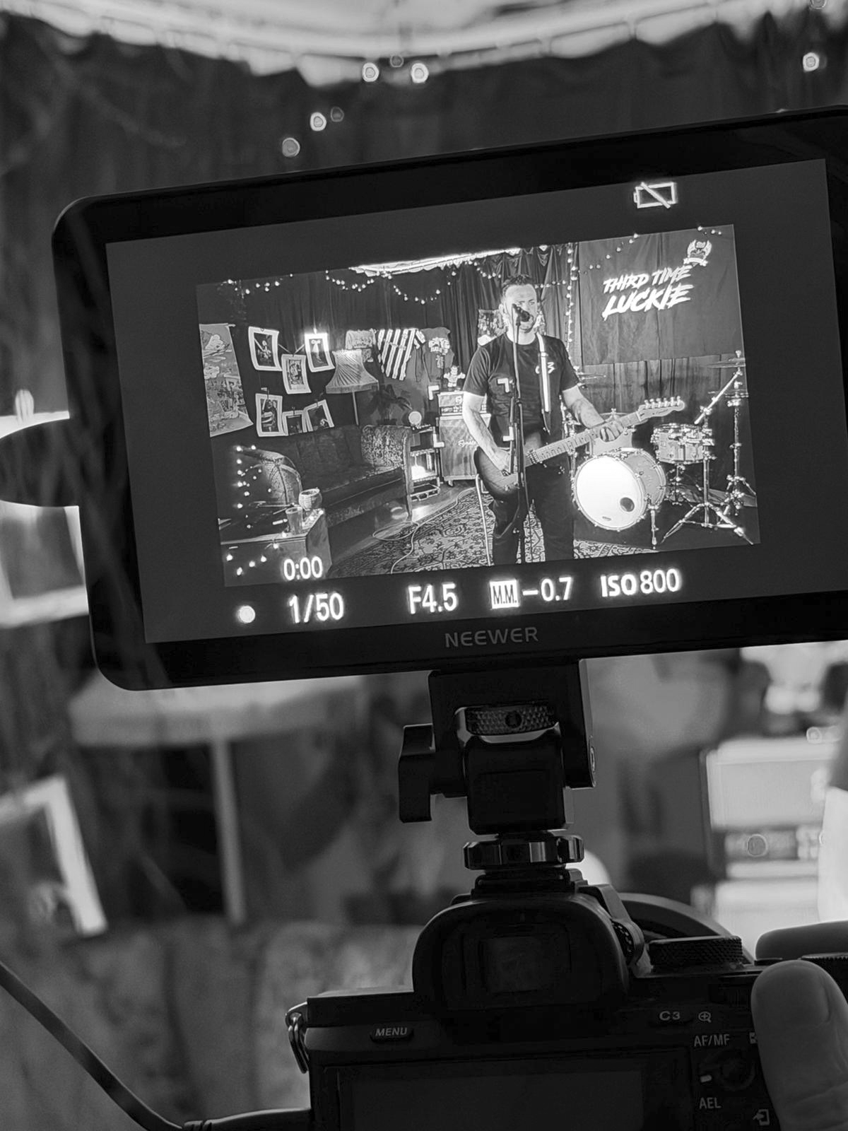 Black and white photo of a camera monitor showing a musician playing electric guitar and singing into a microphone on stage with drums and decorations in the background, and the words "Thank To The Lucky" on the backdrop.