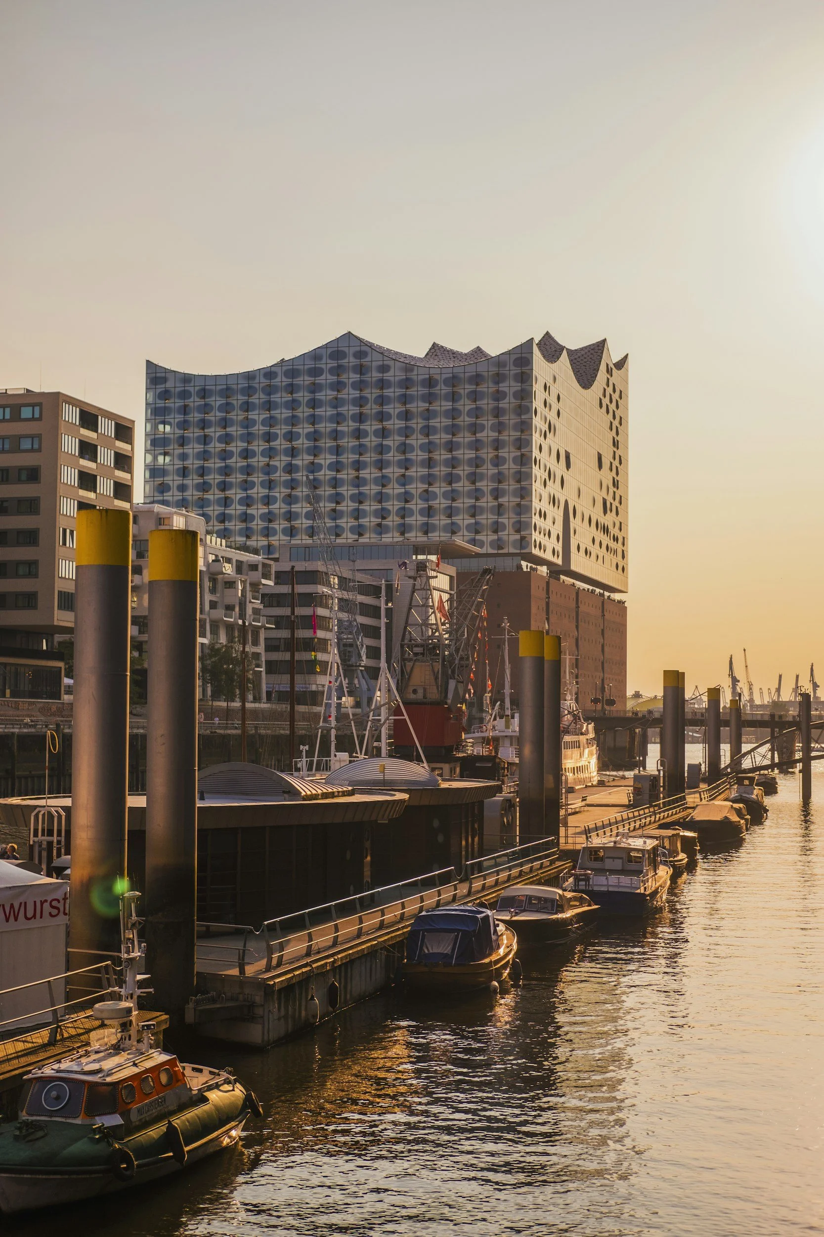 Blick auf einen Hafen bei Sonnenuntergang mit kleinen Booten und modernen Gebäuden, darunter das Elbphilharmonie-Gebäude in Hamburg.