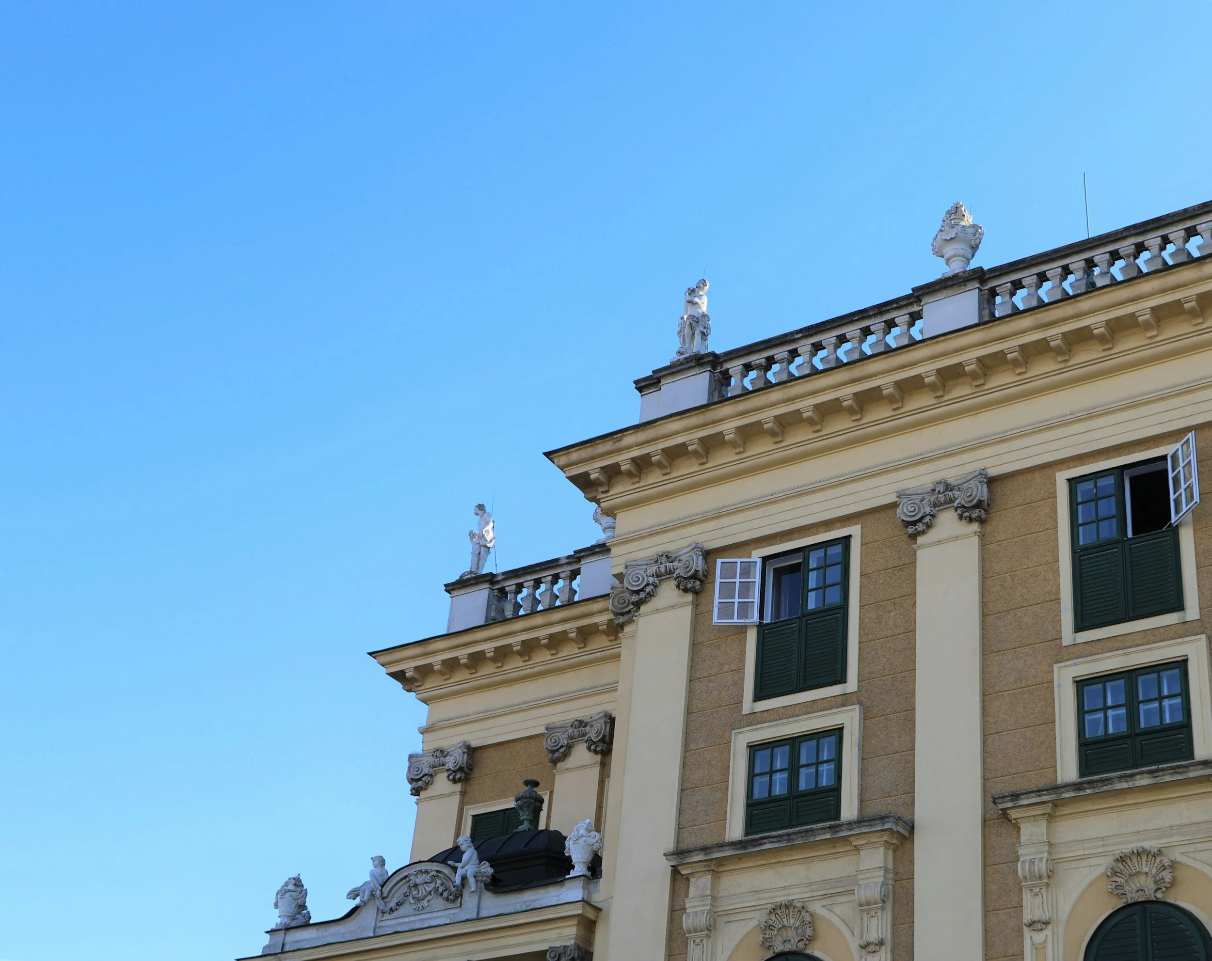 Ansicht eines barocken Gebäudes mit dekorativen Verzierungen, grünen Fenstern und Statuen auf dem Dach, bei klarem blauen Himmel.
